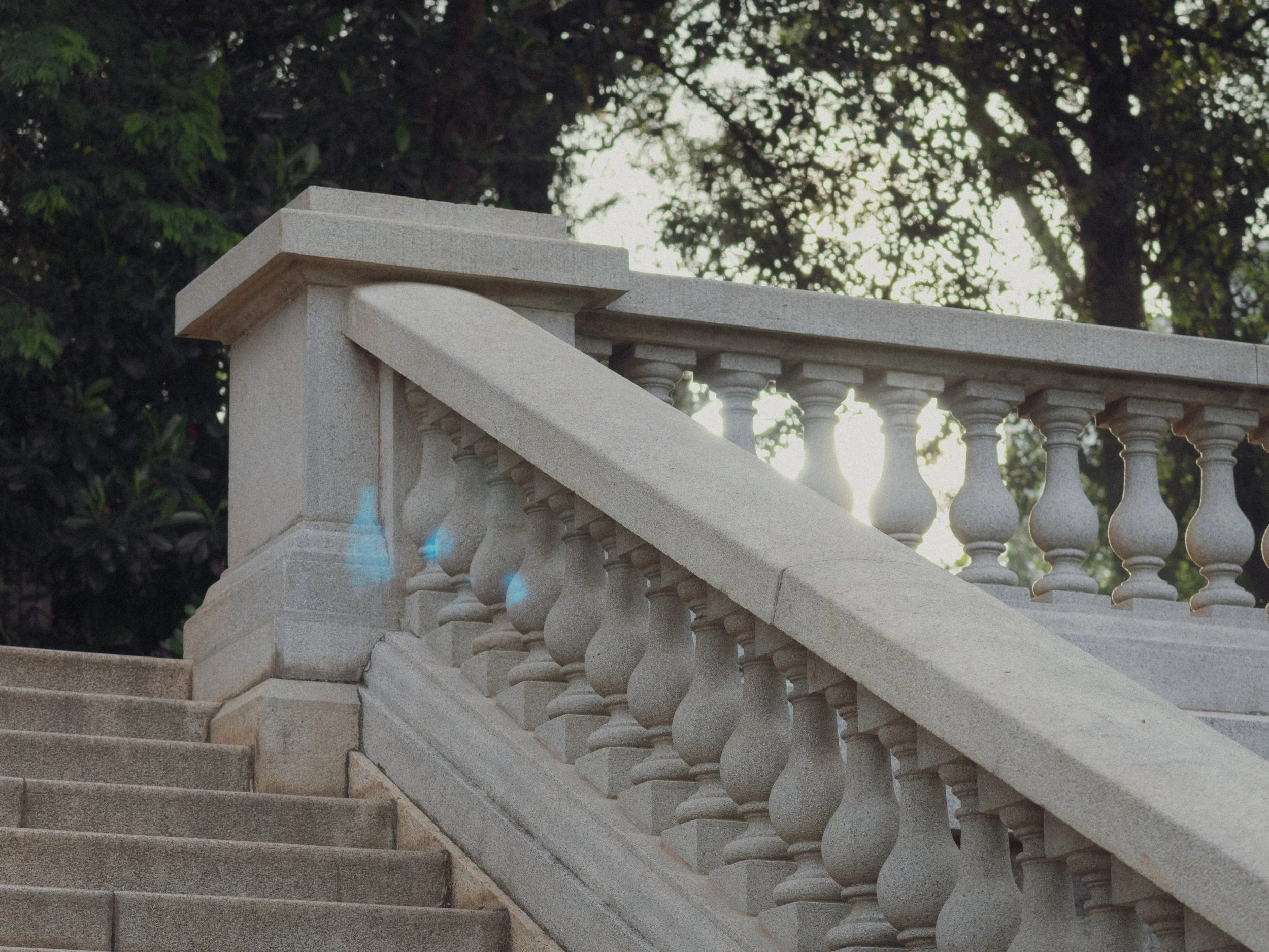 Escadaria de pedra com corrimão de balaustrada, cercada de árvores ao fundo com o sol brilhando por trás.