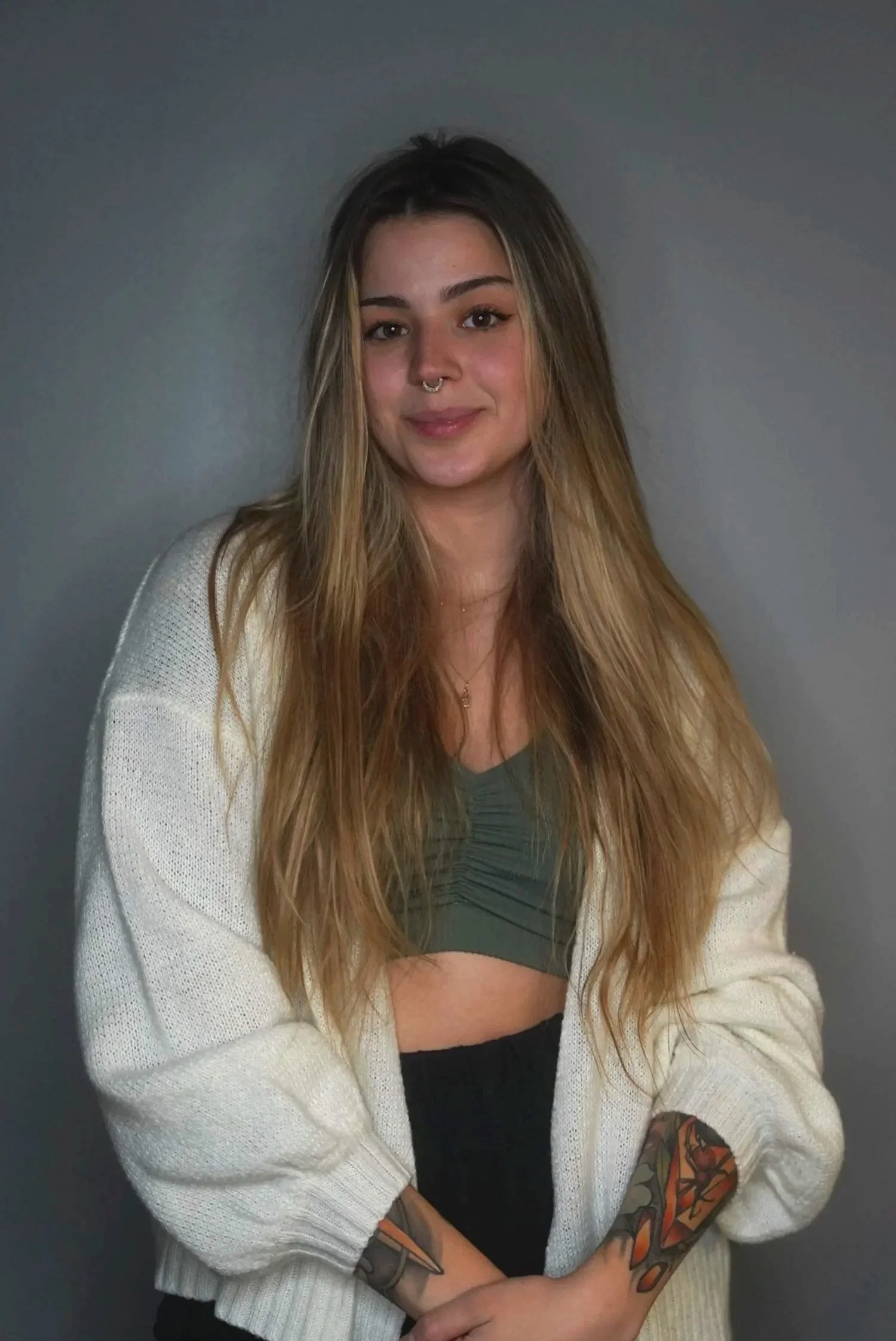 Young woman with long, wavy hair and a septum piercing, wearing a green crop top, a beige cardigan, and black pants, standing against a plain gray wall, with colorful tattoos on her left arm.