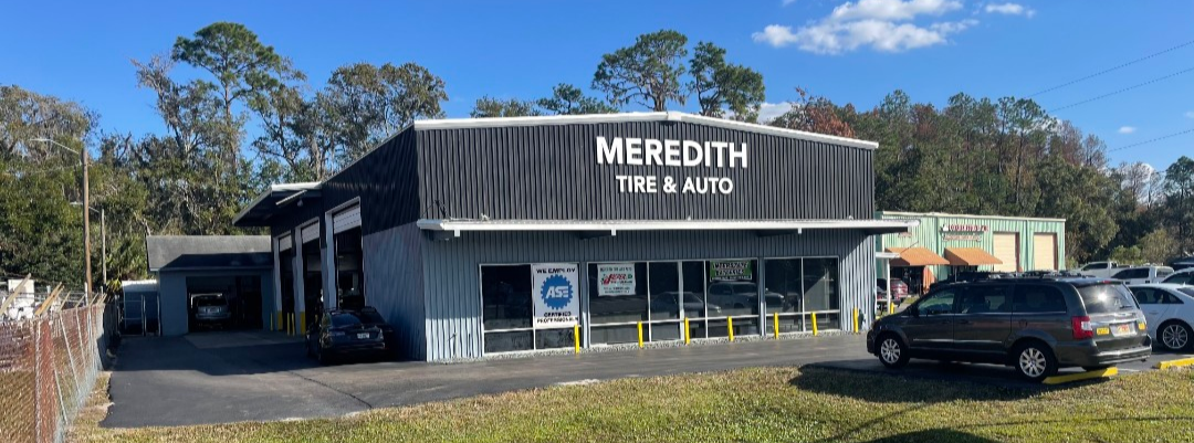 Exterior view of Meredith Tire & Auto shop with a black metal building, large sign, parking lot with cars, and trees in the background.