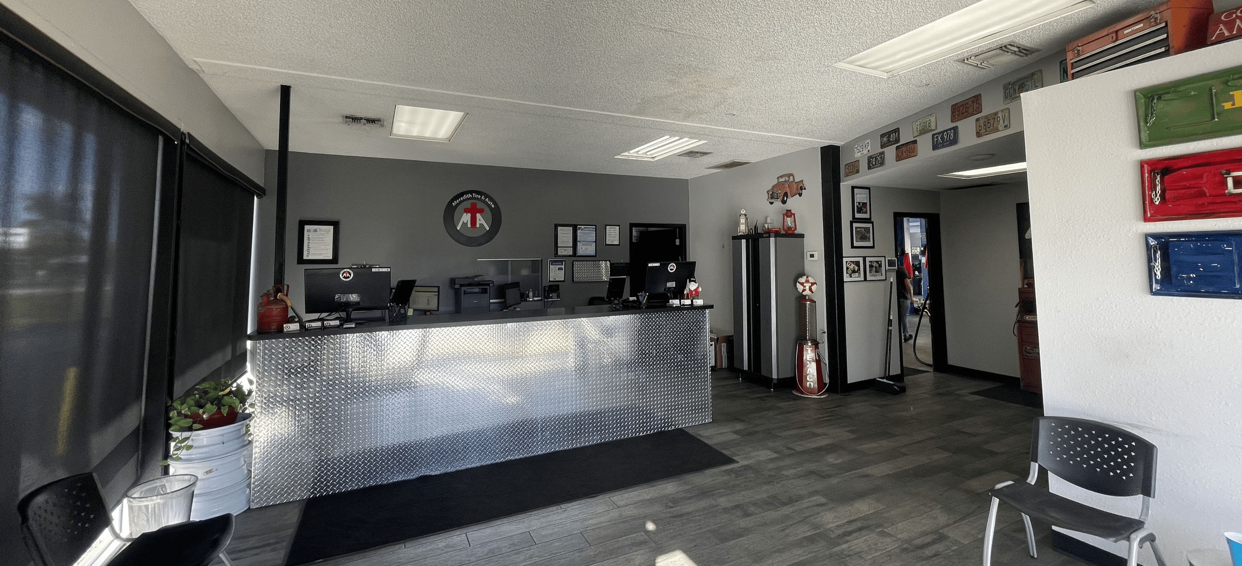 Car service center front desk area with a metallic counter, computer monitors, wall decor with license plates, and a waiting chair.