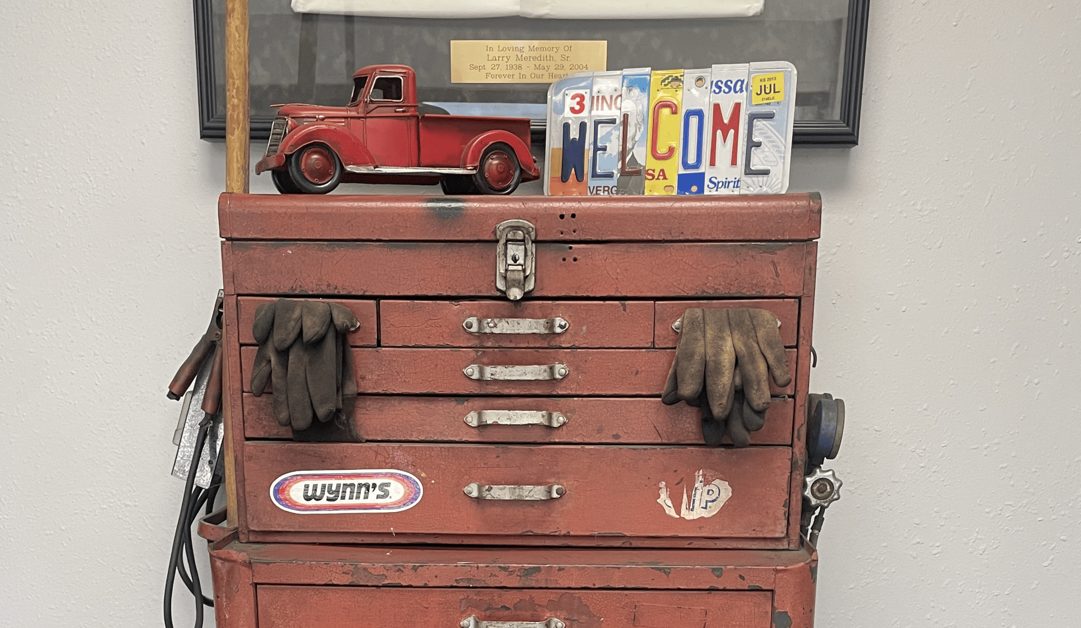 Vintage red tool chest with gloves hanging on handles, a small red toy truck on top, and decorative welcome messages and license plates behind it.