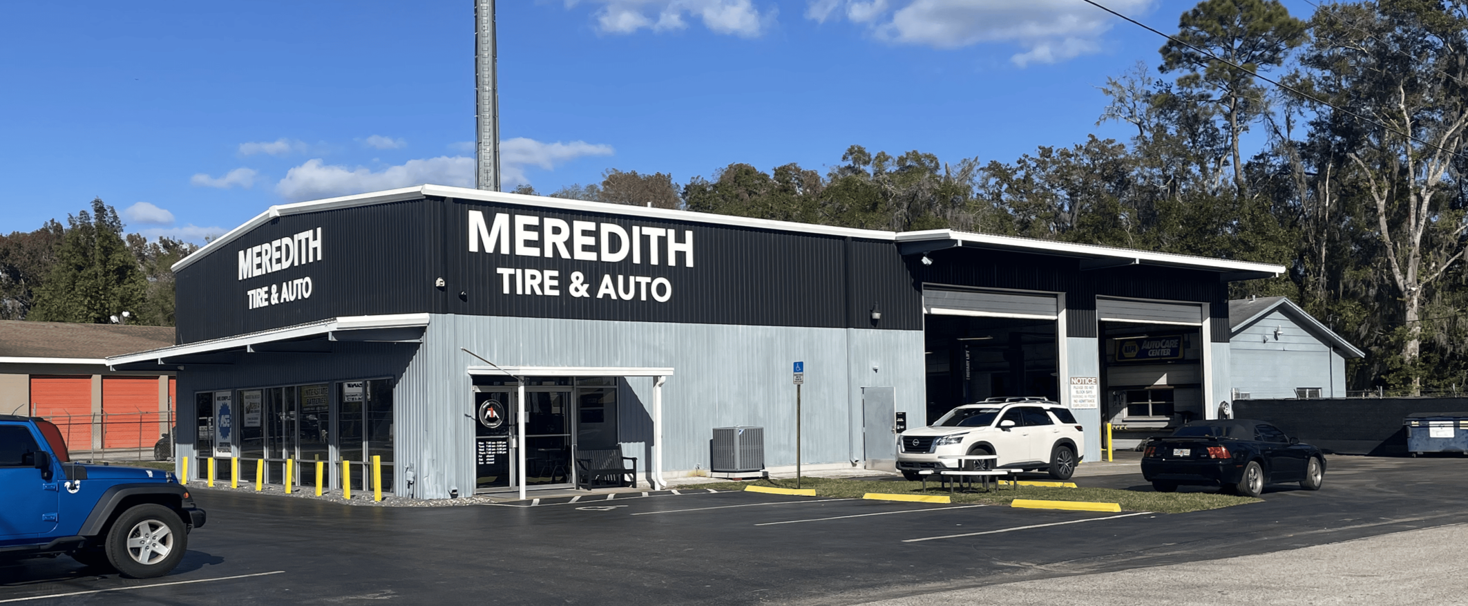 Exterior view of Meredith Tire & Auto shop with a black metal building, white signage, and several parked cars including a white SUV and a black sedan. Clear blue sky and trees in the background.