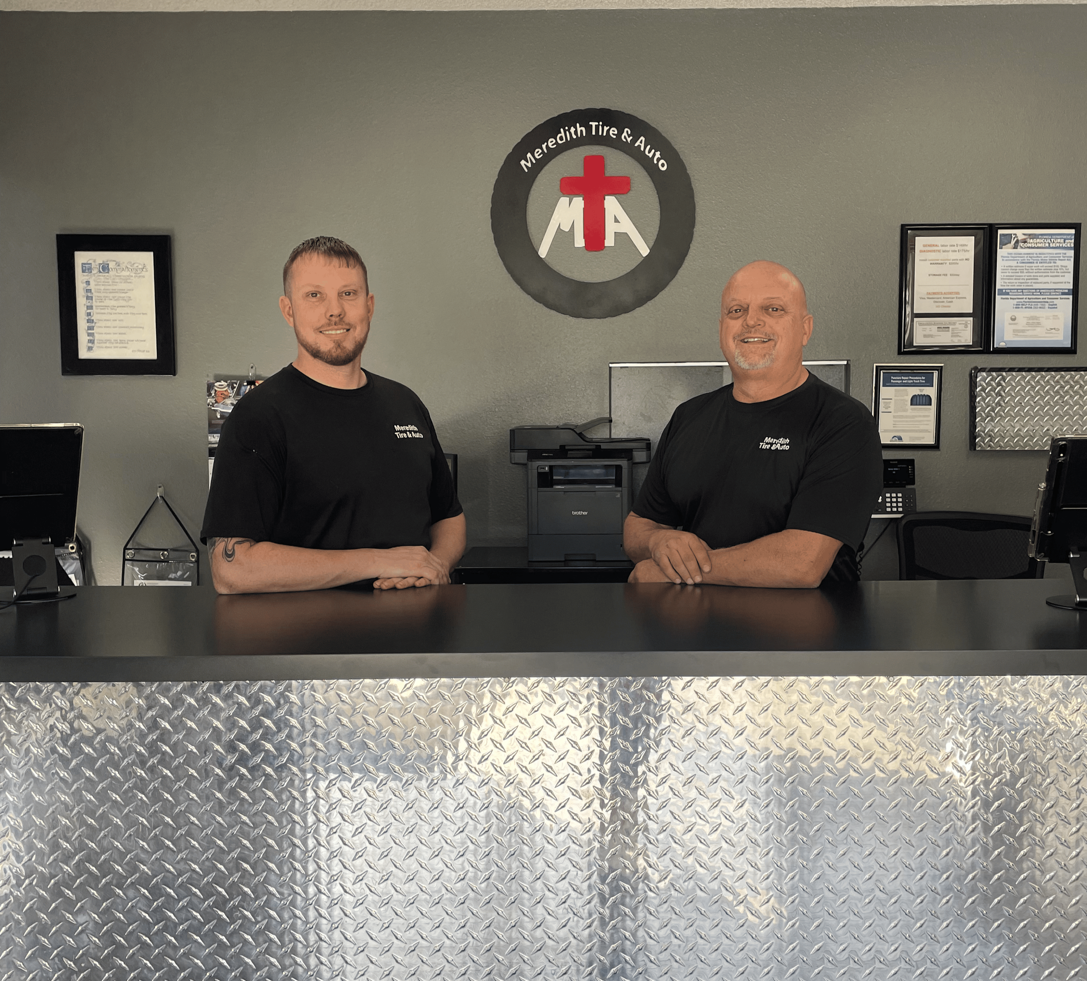 Two men standing behind a desk at Meredith Tire & Auto, smiling. The logo is on the wall behind them, with certificates and office equipment visible.