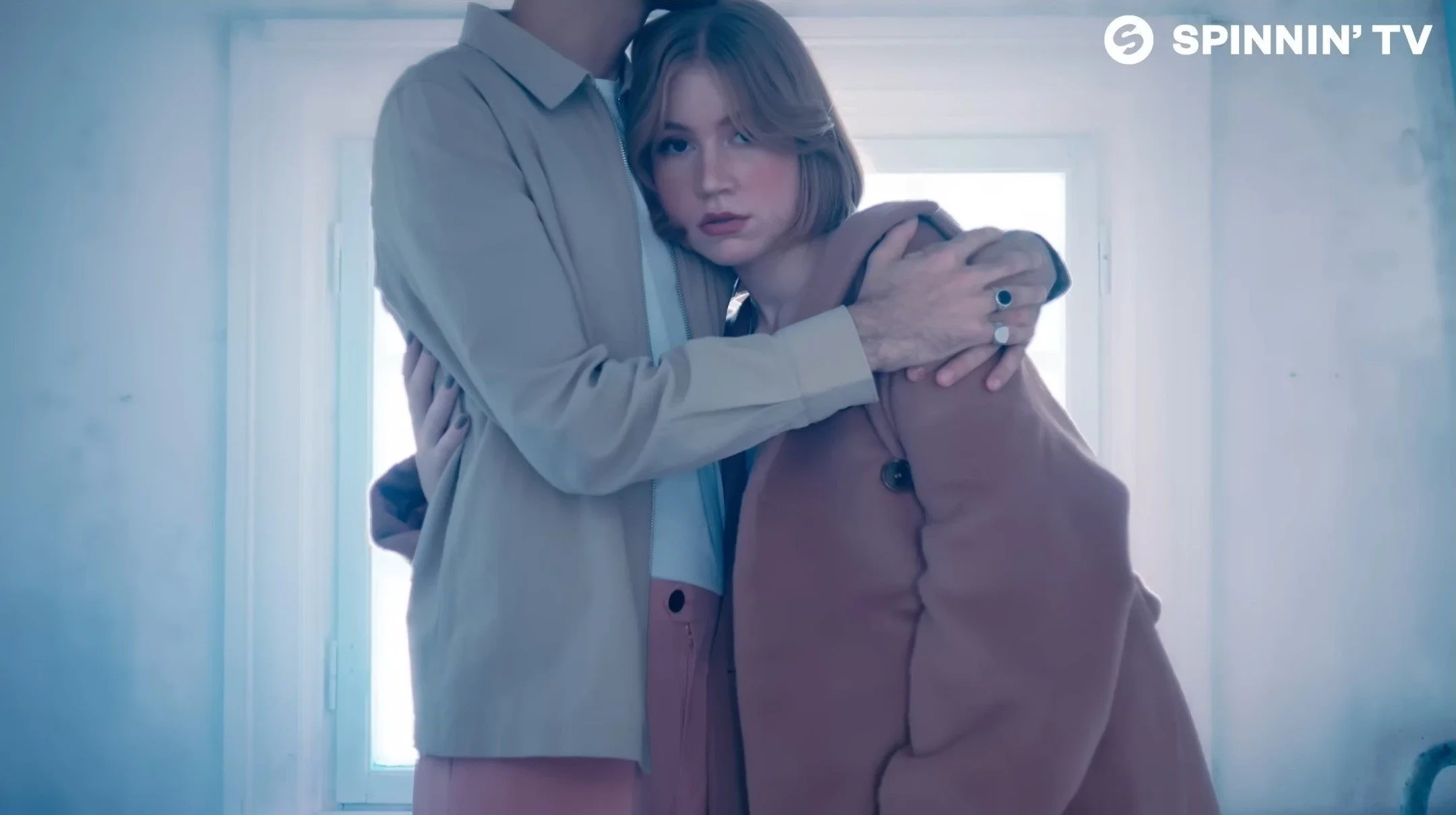 A young woman with short blonde hair and a serious expression, standing close to an older man, with her head resting on his chest. The man is holding her with both arms, and they are standing in front of a window that lets in bright light. The overall mood appears emotional or introspective.