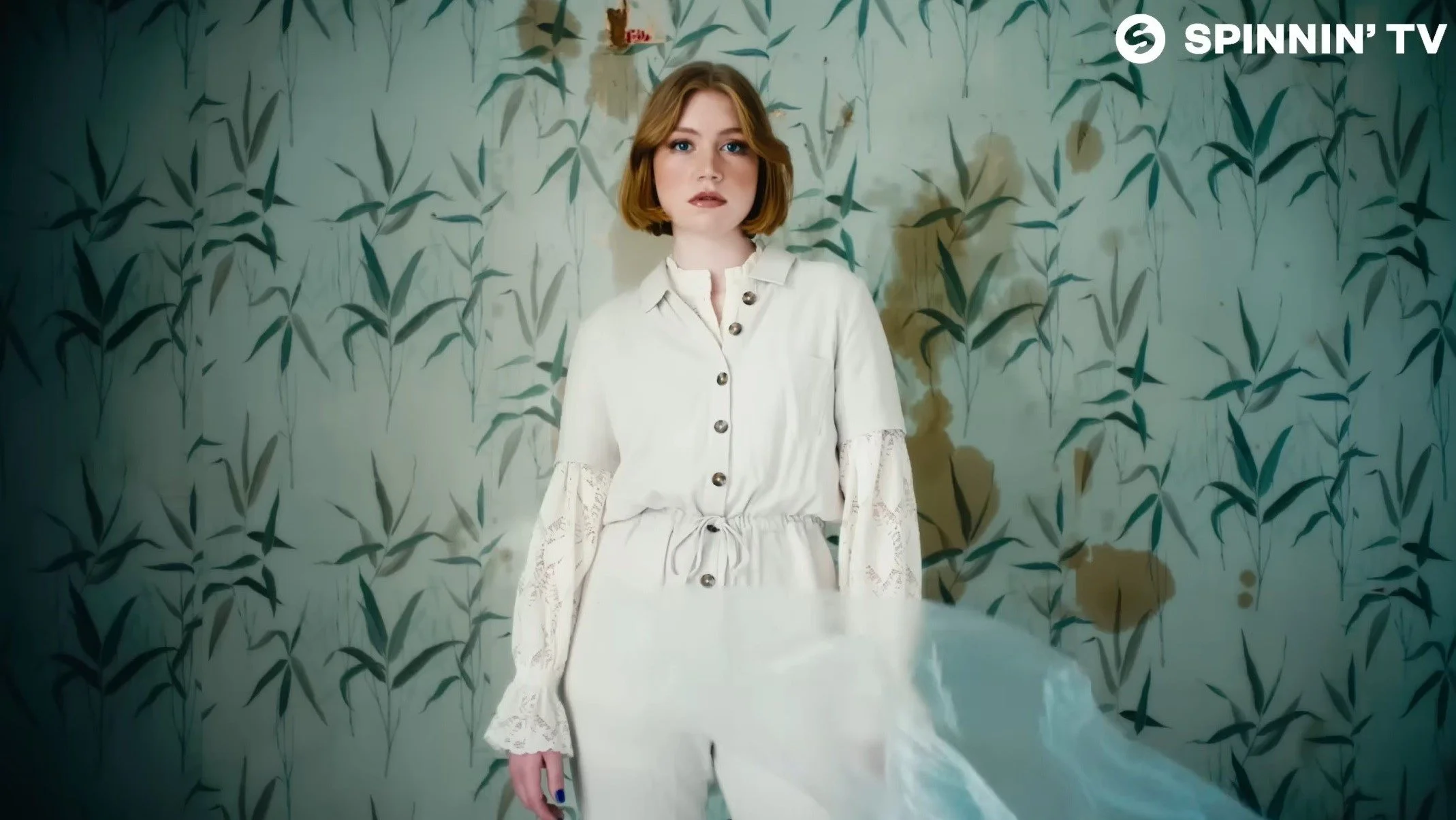 A woman with short, wavy red hair wearing a white dress with lace sleeves and buttons stands in front of a green leafy patterned wall.