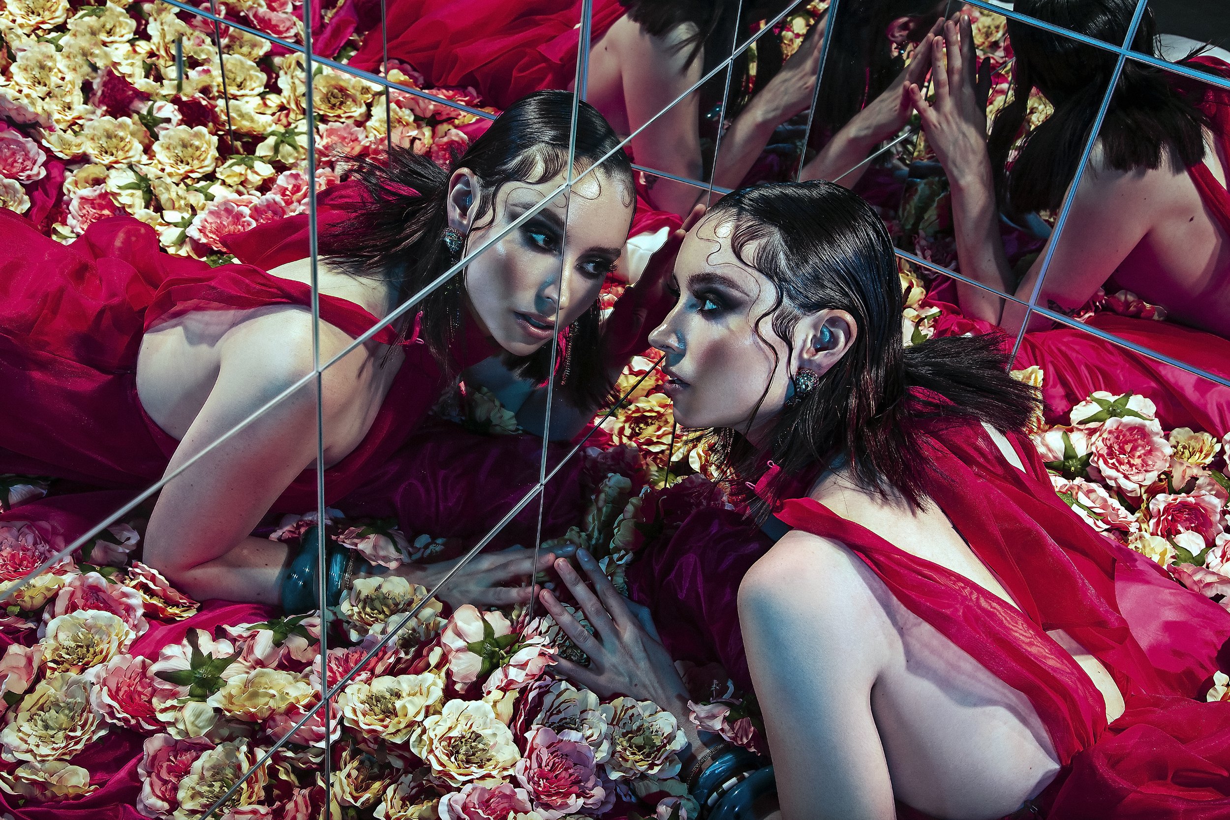 Two women with dark hair and dramatic makeup lying among pink, peach, and cream flowers, looking into a mirror with multiple reflections.