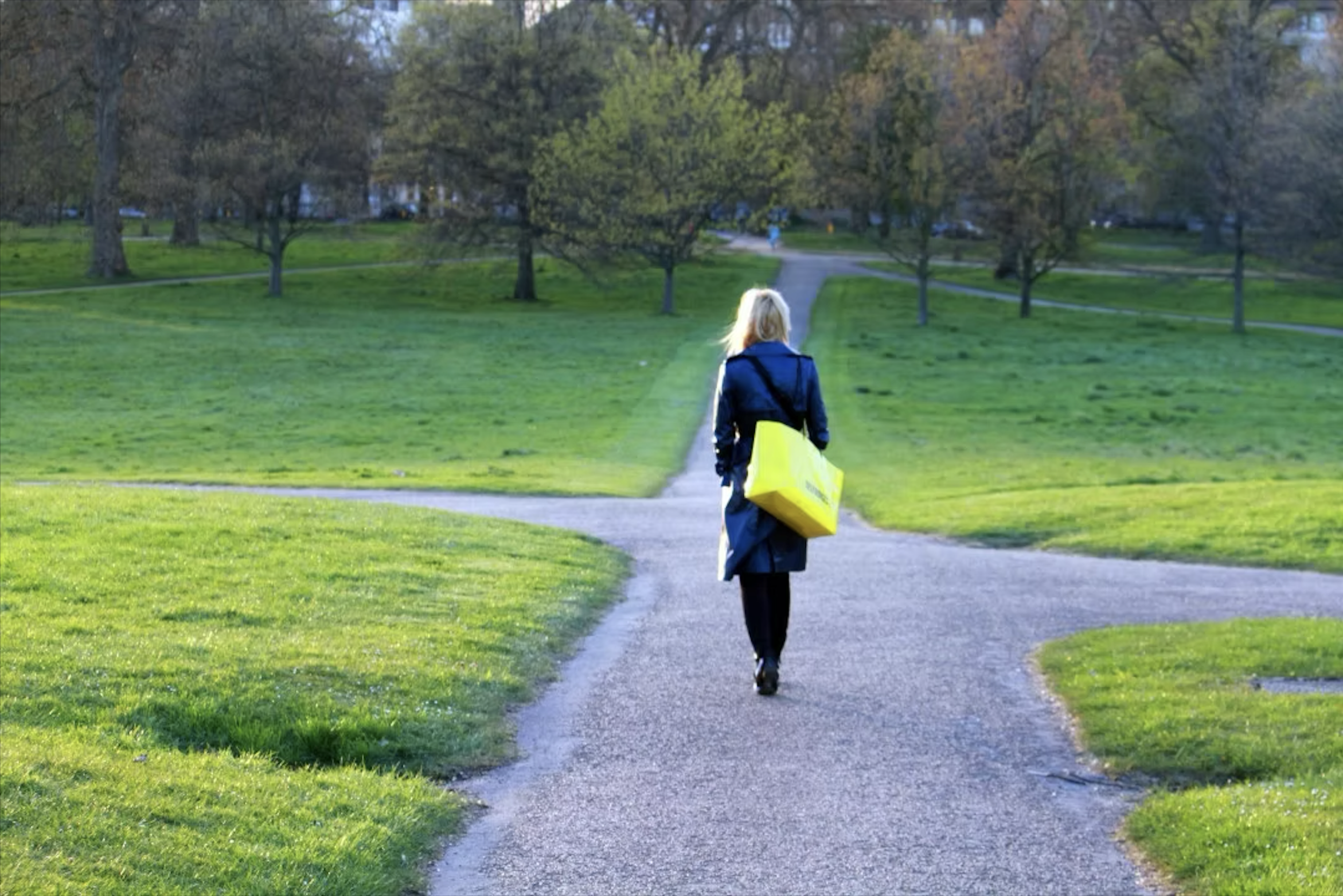 A woman with blonde hair walking on a winding park pathway surrounded by green grass and trees. She is carrying a large yellow bag and dressed in a coat.