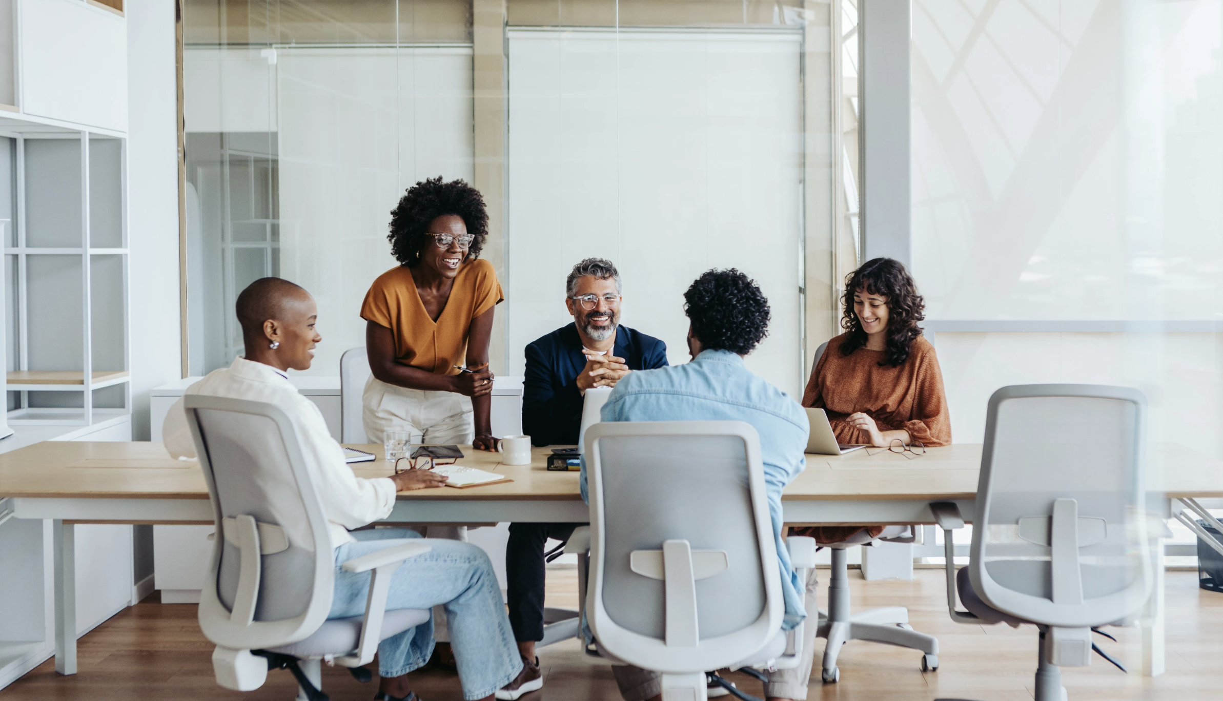 A diverse group of five people having a meeting in a modern office conference room, with a woman standing and smiling while others sit around the table, some with laptops and notebooks.