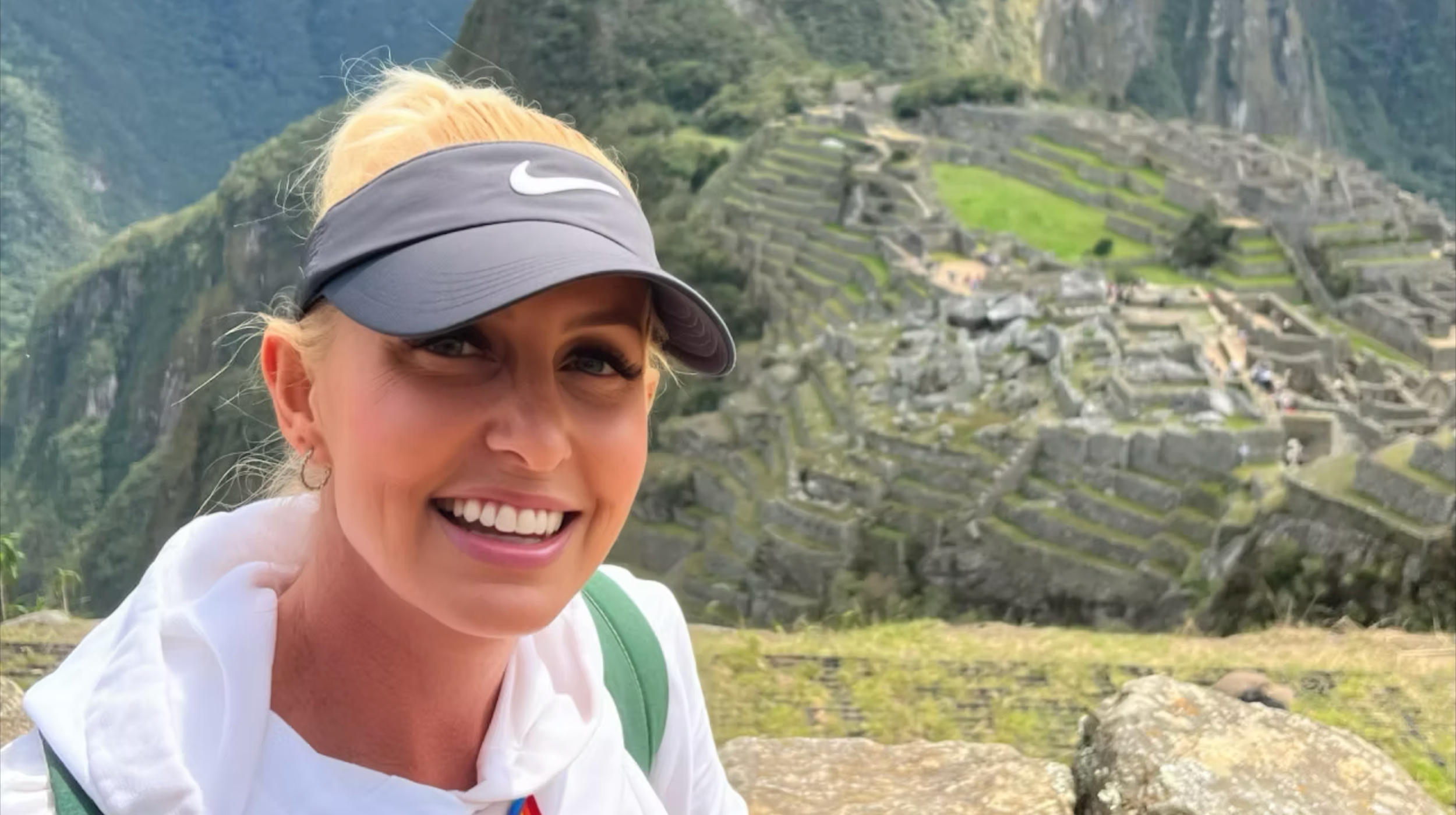 A smiling woman wearing a gray Nike visor and white jacket takes a selfie with Machu Picchu ruins and green mountains in the background.