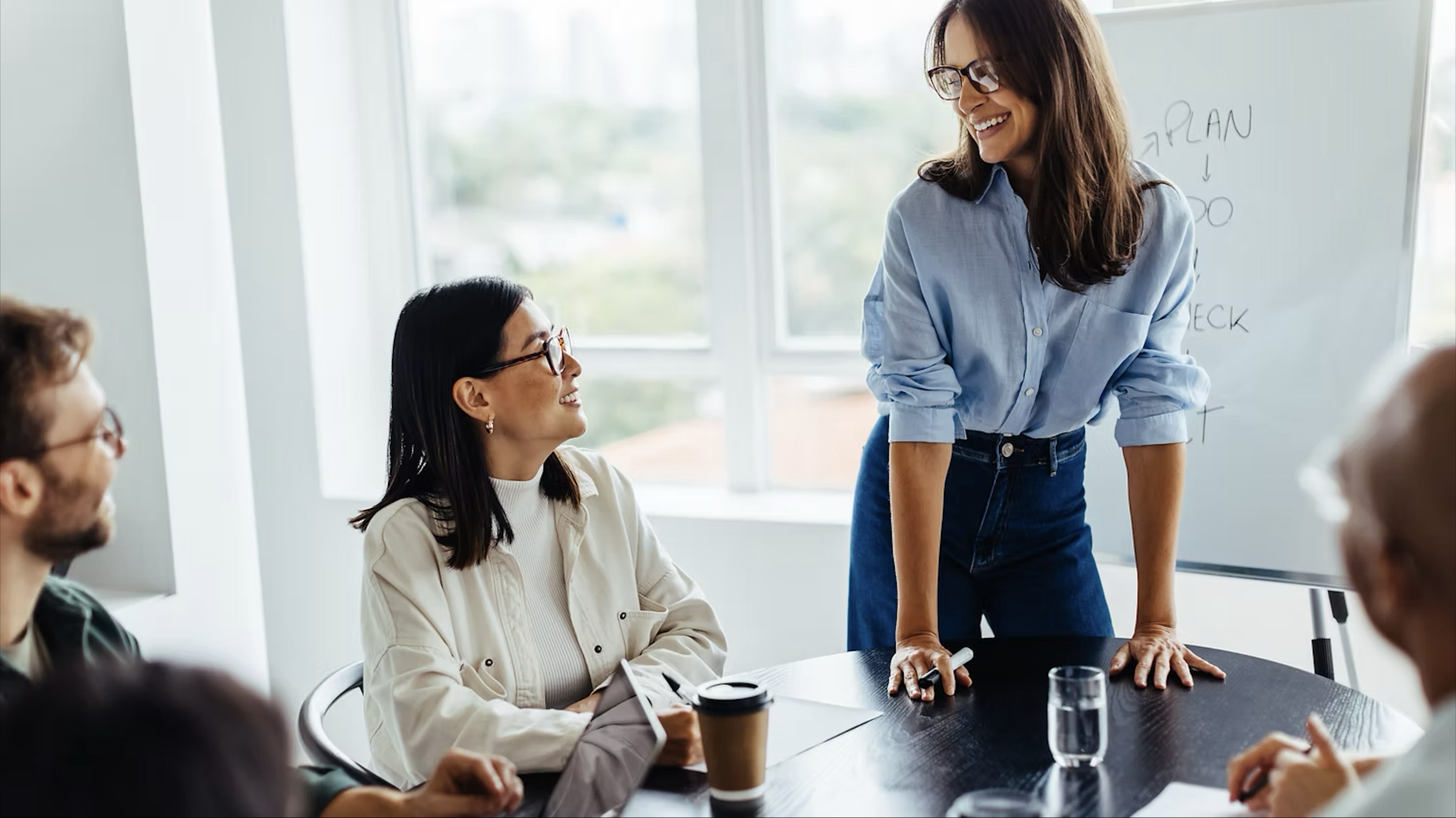 A woman standing and leaning on a table, smiling and speaking to a group of people sitting around the table during a meeting in a bright room with windows.