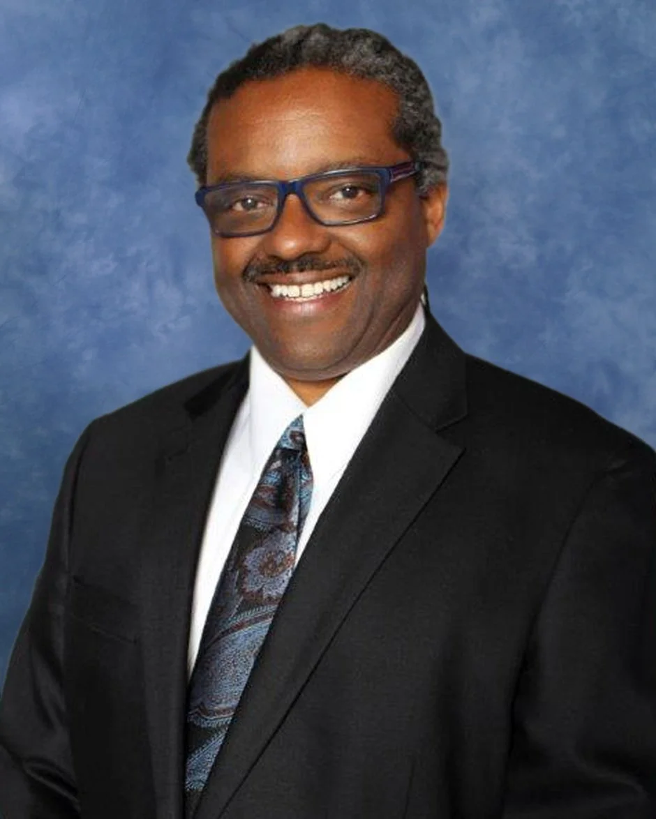 A man wearing a black suit, white shirt, and a colorful tie, smiling at the camera against a blue background.