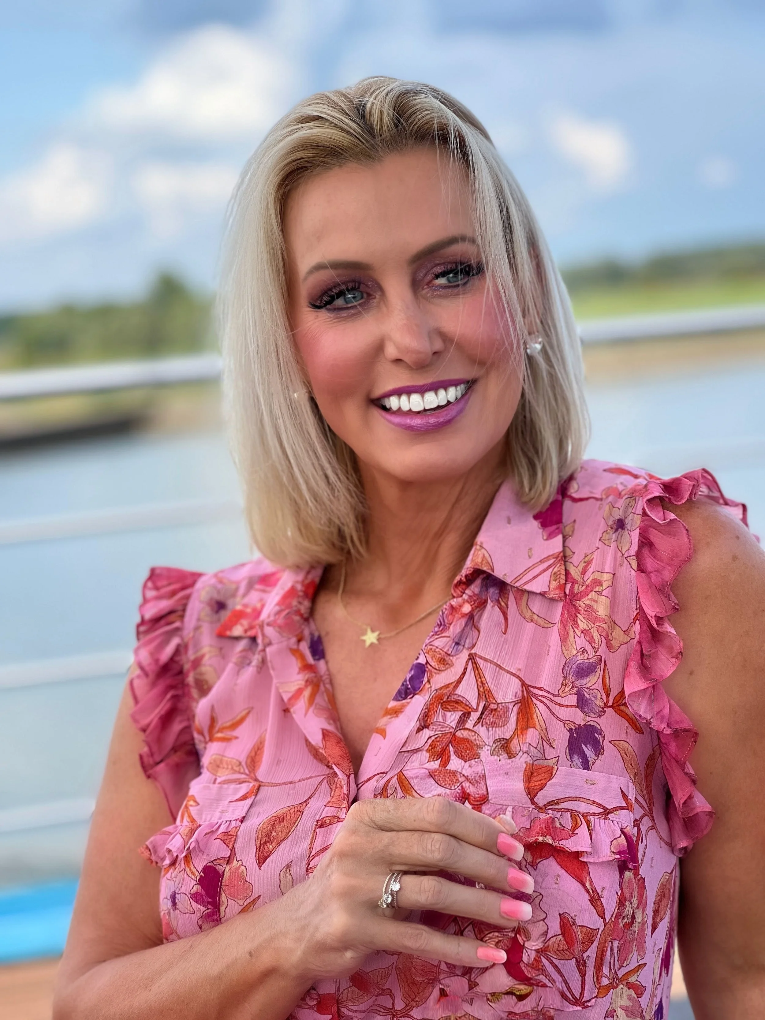 A smiling woman with blonde hair wearing a pink floral sleeveless top, jewelry, and makeup, posing outdoors with a body of water and sky in the background.