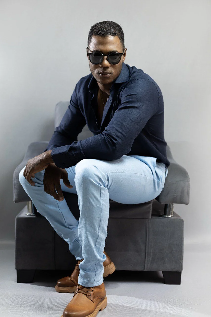 Hombre con gafas de sol, camiseta azul oscuro, pantalones claros, y botas marrones, sentado en un sillón gris contra una pared blanca.