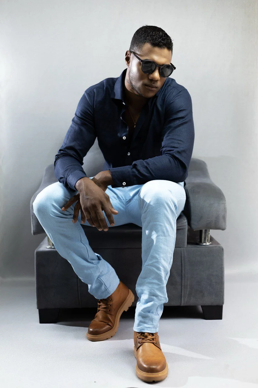 Hombre joven con gafas de sol, camiseta azul y pantalones claros, sentado en un sillón gris, usando botas marrones.