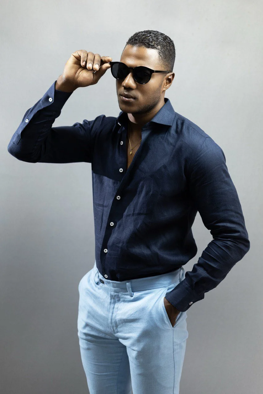 Hombre joven con piel oscura usando gafas de sol negras, camisa azul y pantalones claros, se acomoda las gafas con la mano derecha y mira de lado, con una expresión confiada.