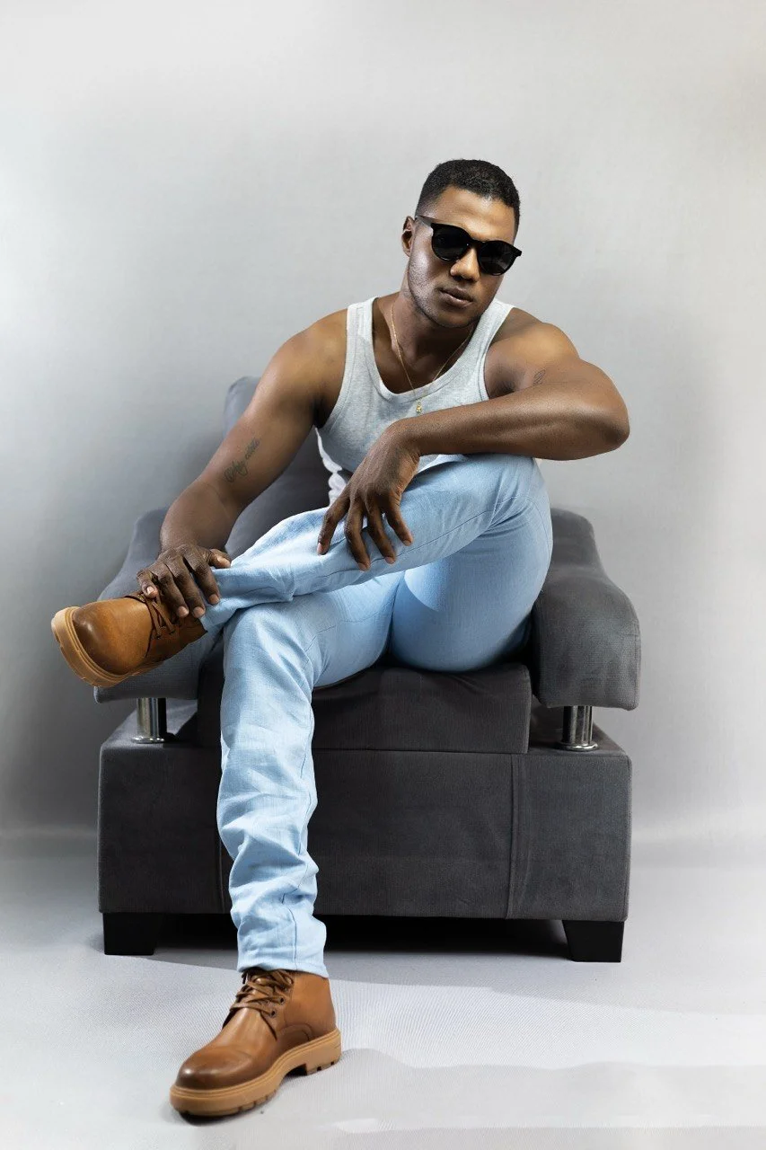 Joven hombre sentado en un sillón con gafas de sol, camisa sin mangas gris, pantalones claros y botas marrones, en un fondo gris liso.