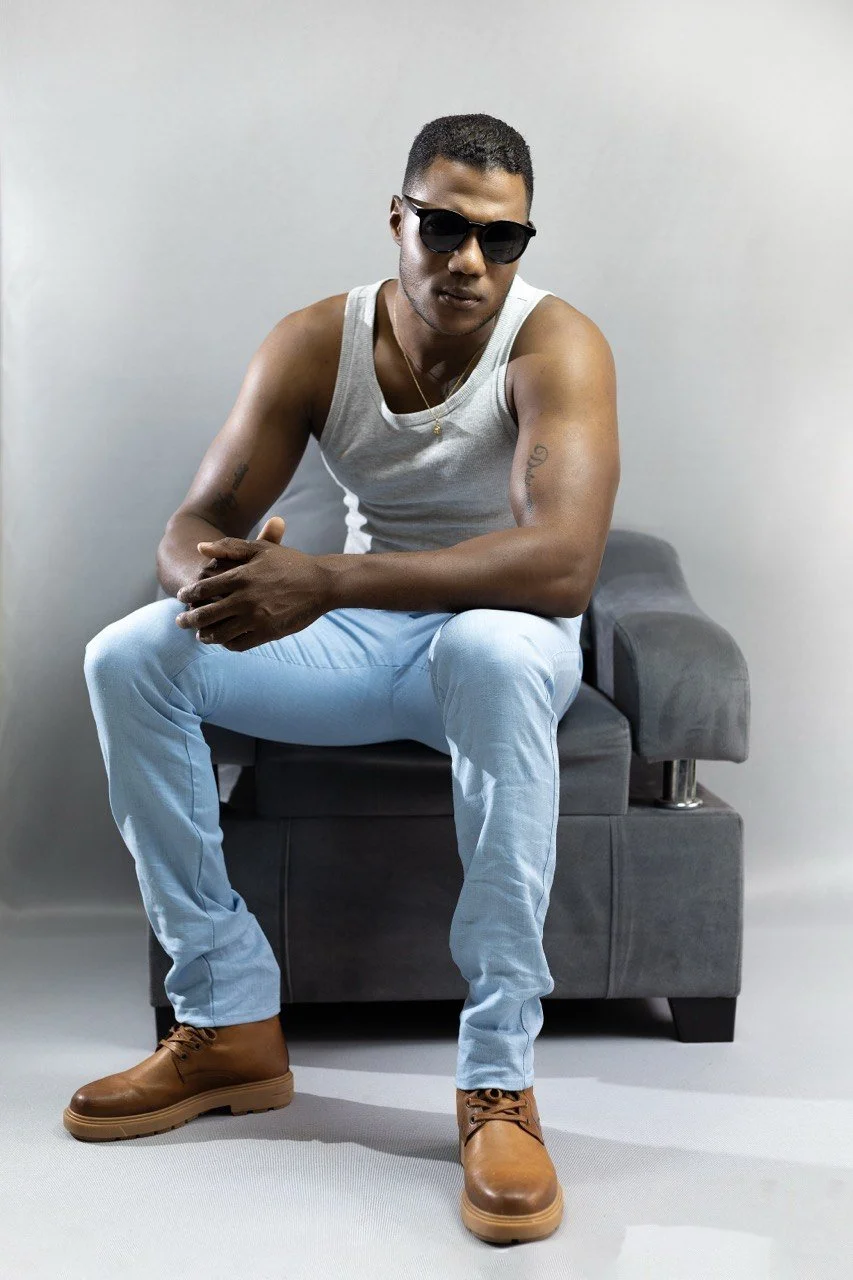 Joven con piel oscura, gafas negras, camiseta sin mangas gris, pantalones claros y botas marrones sentado en un sillón gris, con una expresión confiada.