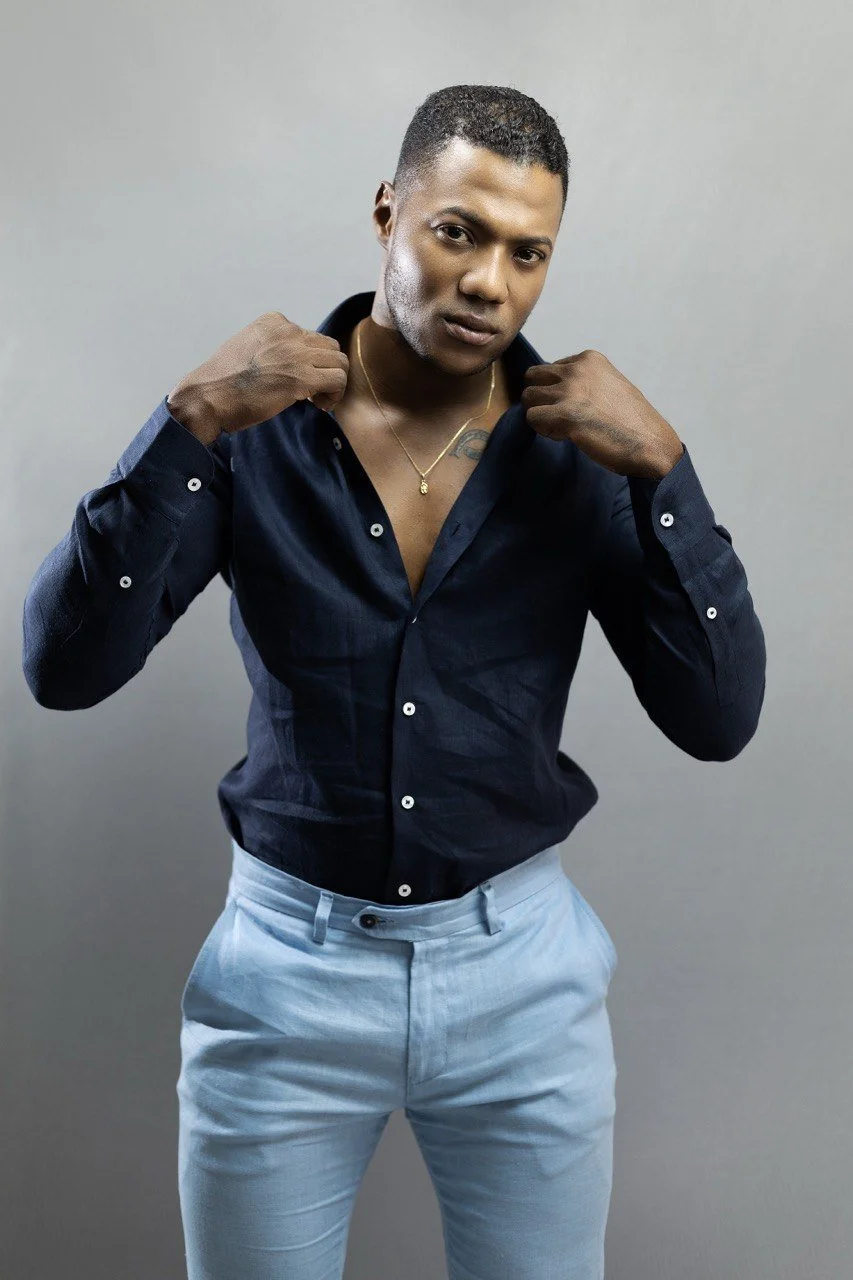 Joven hombre con piel oscura, cabello corto, usando camisa azul oscuro y pantalones claros, ajustándose la camisa y mirando a la cámara.