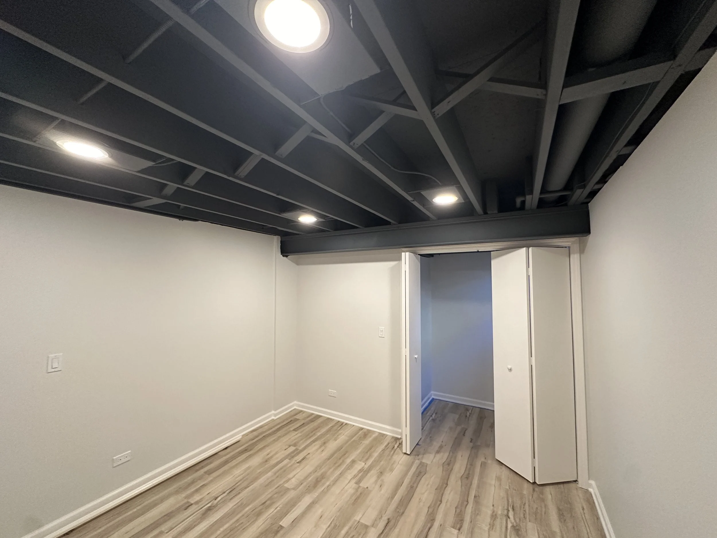 Empty room with white walls, wood flooring, and an open closet with sliding doors. The ceiling is black with exposed beams and recessed lighting.
