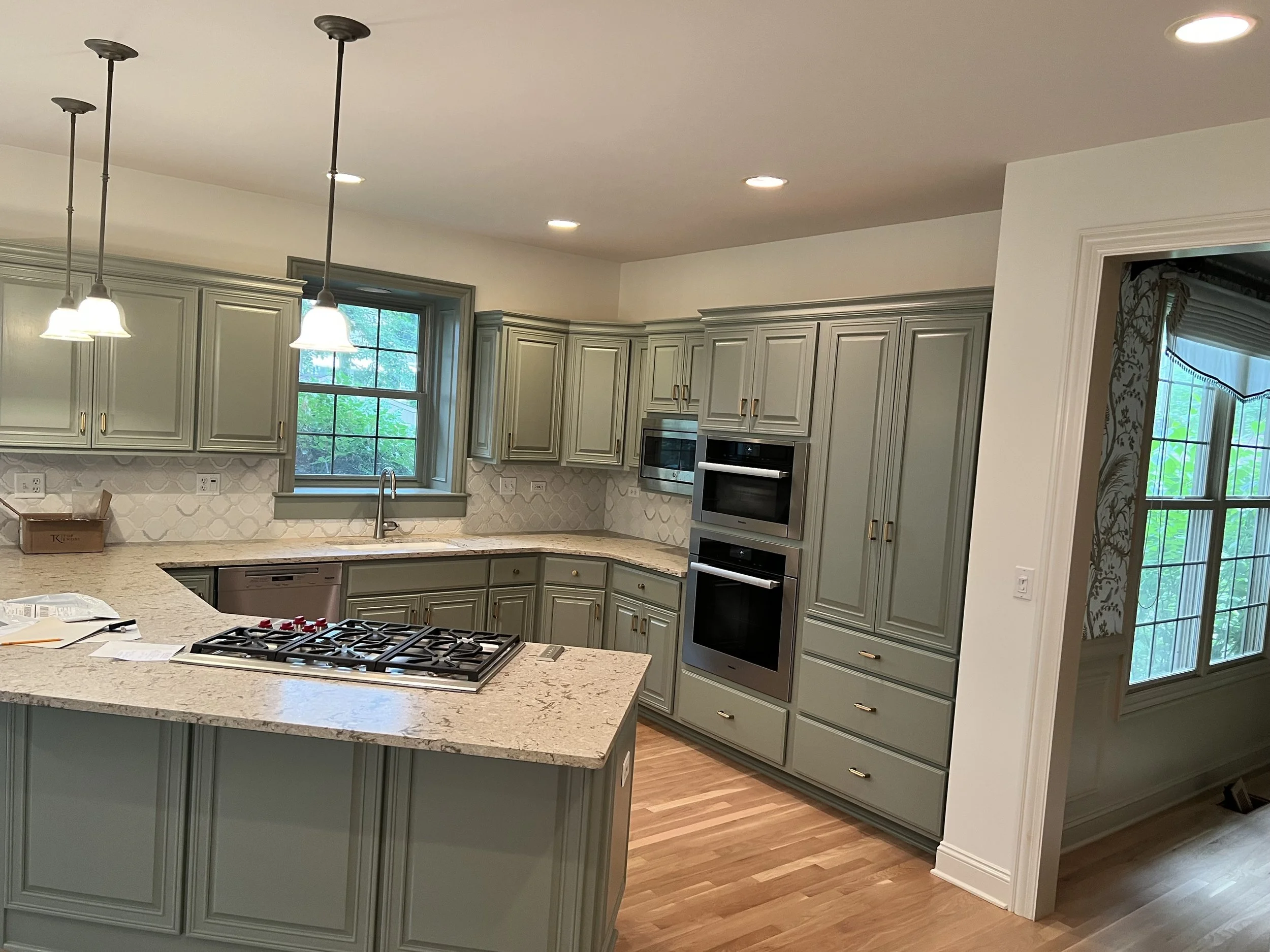 Kitchen with green cabinetry, granite countertops, a window above the sink, built-in microwave and oven, pendant lights, and hardwood flooring.