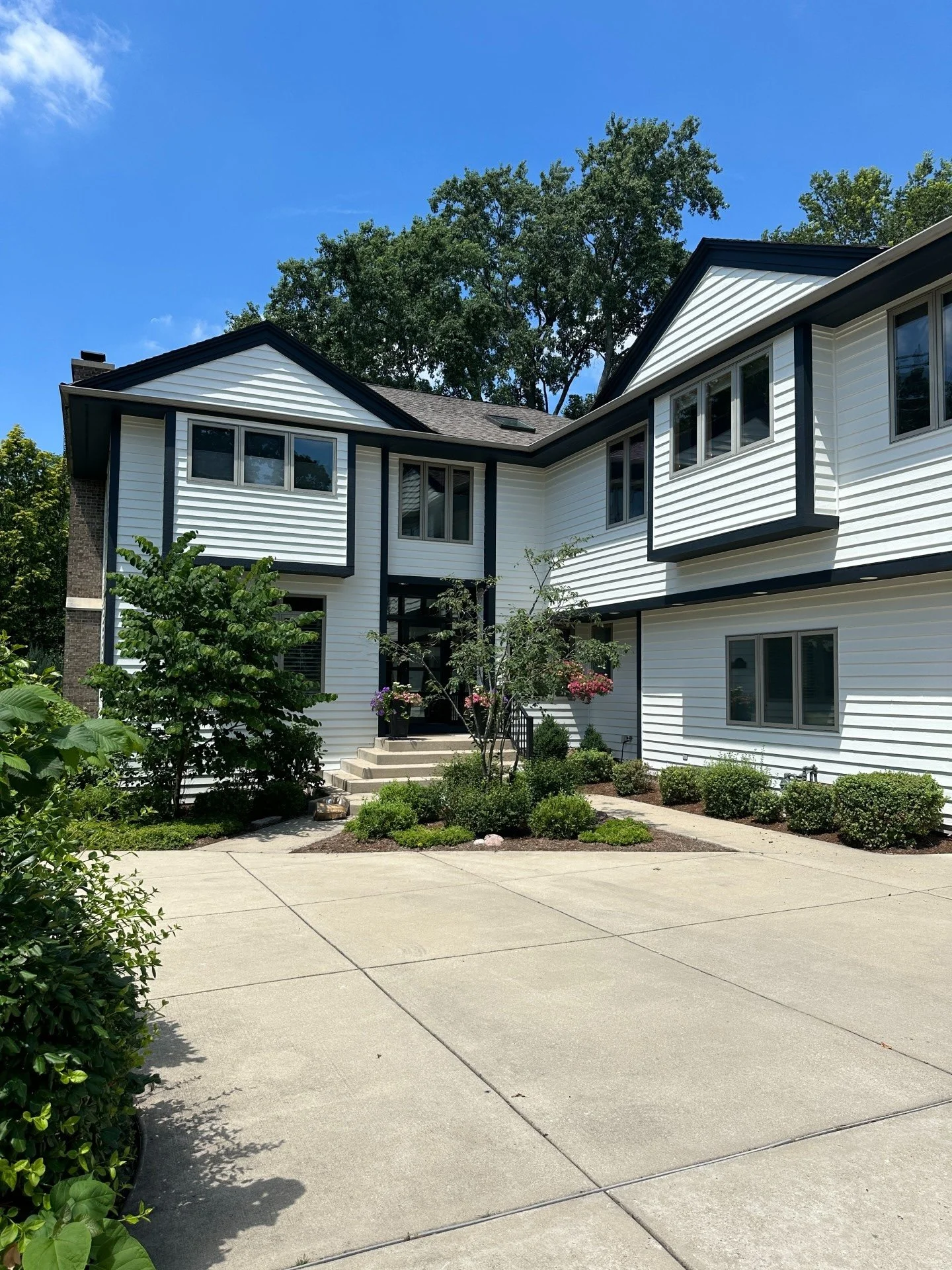 A modern two-story white house with black trim, surrounded by greenery, with a concrete driveway leading up to the front steps.