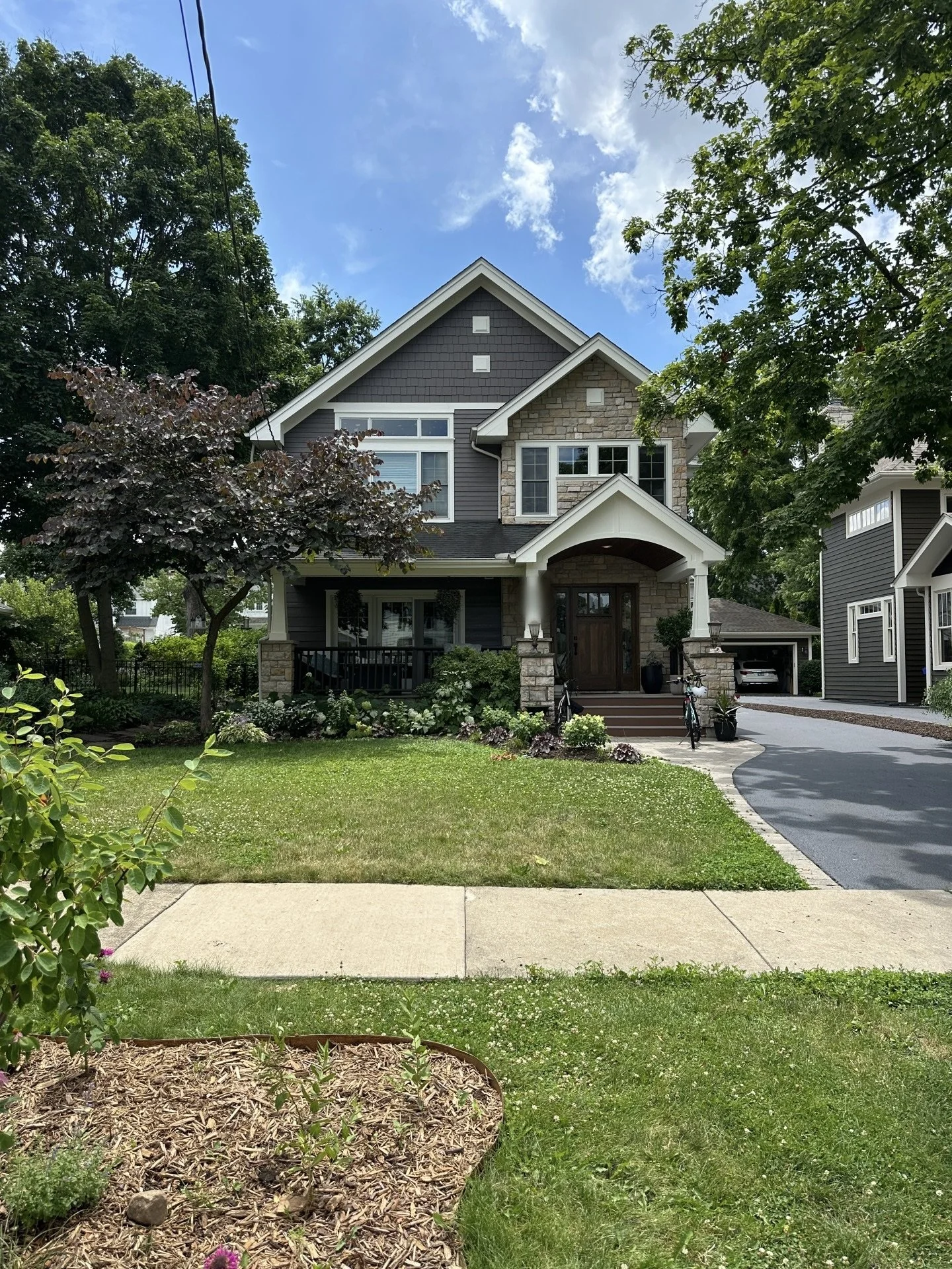 A two-story house with gray siding, stone accents, and a gabled roof. The house has a front porch with steps leading up to a wooden door, and a driveway to the right. There are green trees and a well-maintained lawn with flower beds in front.