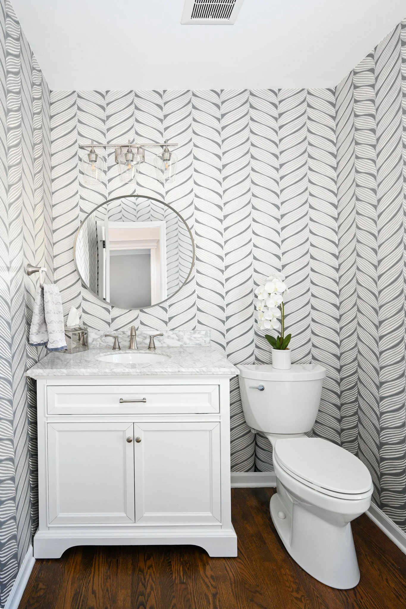 A small bathroom with geometric patterned wallpaper, a white vanity with a marble top, and a white toilet with a potted orchid on top.