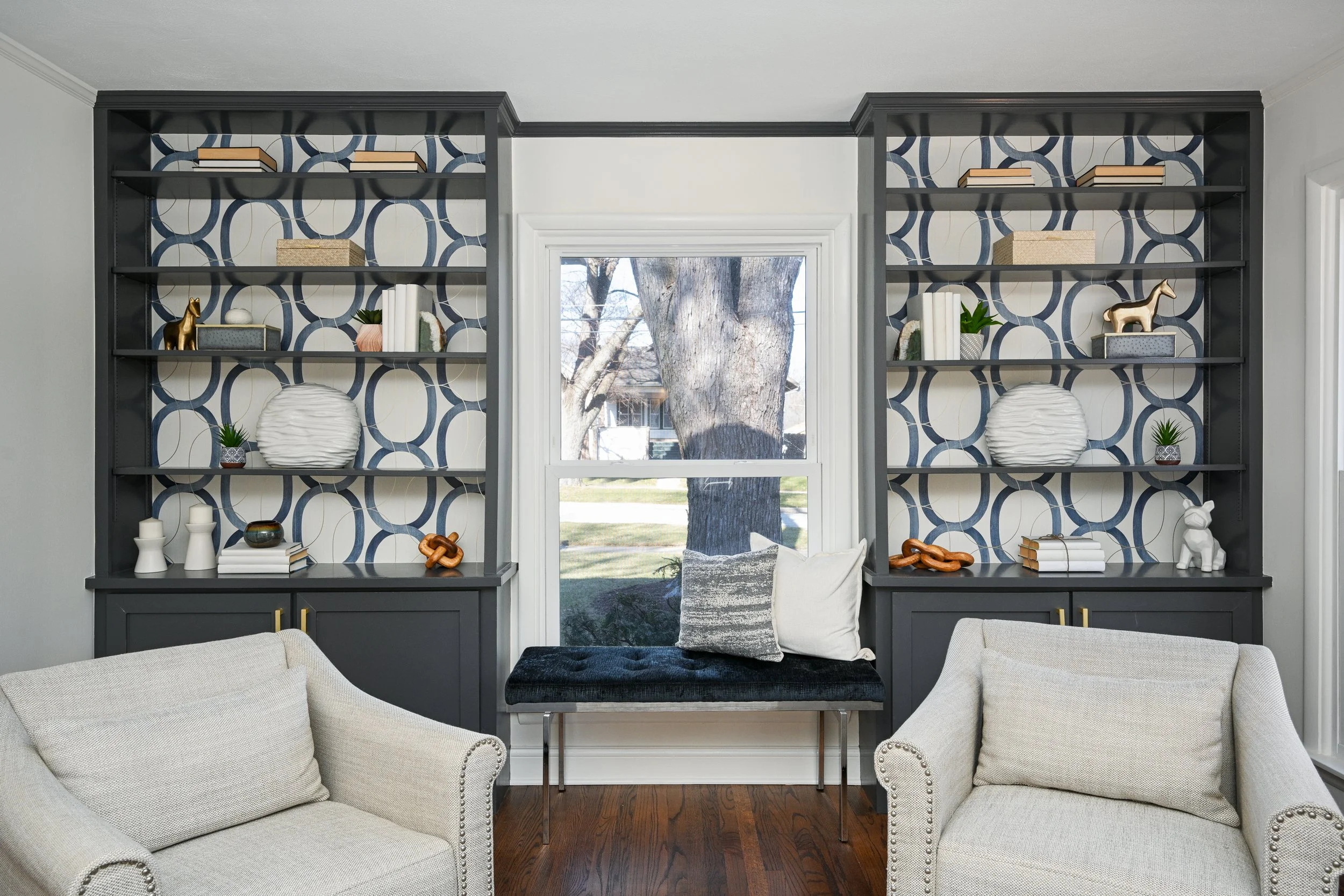 Living room with built-in dark gray bookshelves on both sides of a window, decorated with books, vases, and decorative objects, beige armchairs, and a window seat with cushions, hardwood floors.