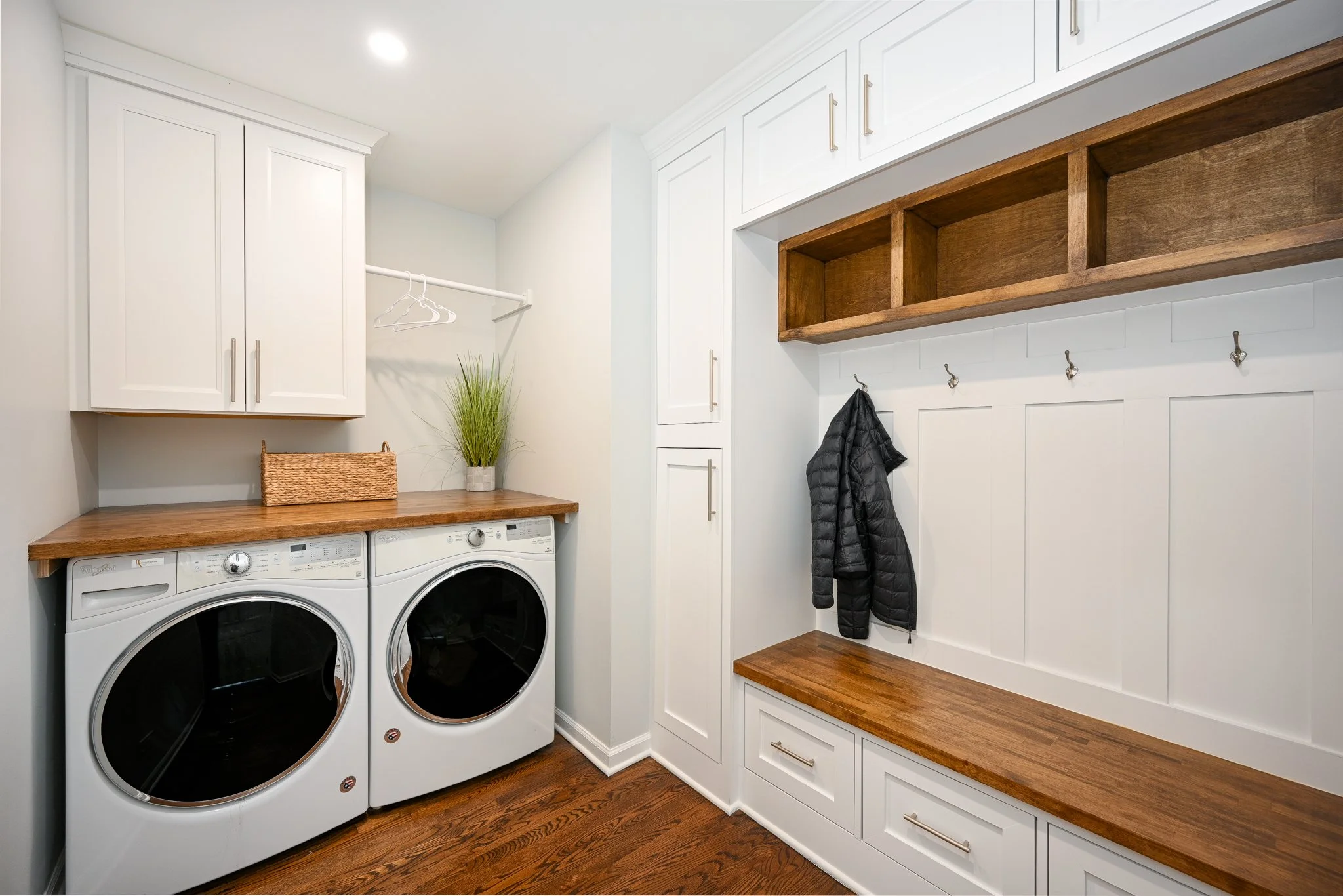 A laundry room with white cabinets, a wooden countertop, and a washer and dryer. There are coat hooks on the wall, a black jacket hanging, a bench, and a small plant.