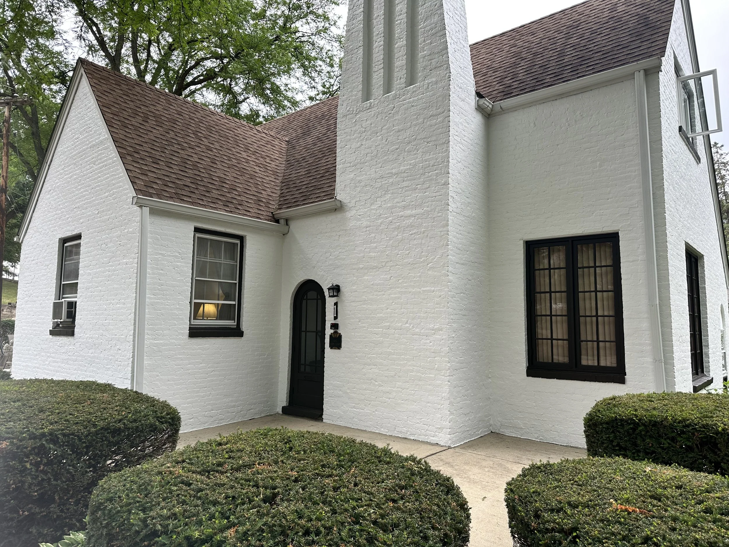 White brick house with black window frames, black front door, small front porch, bushes in front, and a chimney on the side with red shingle roofing.
