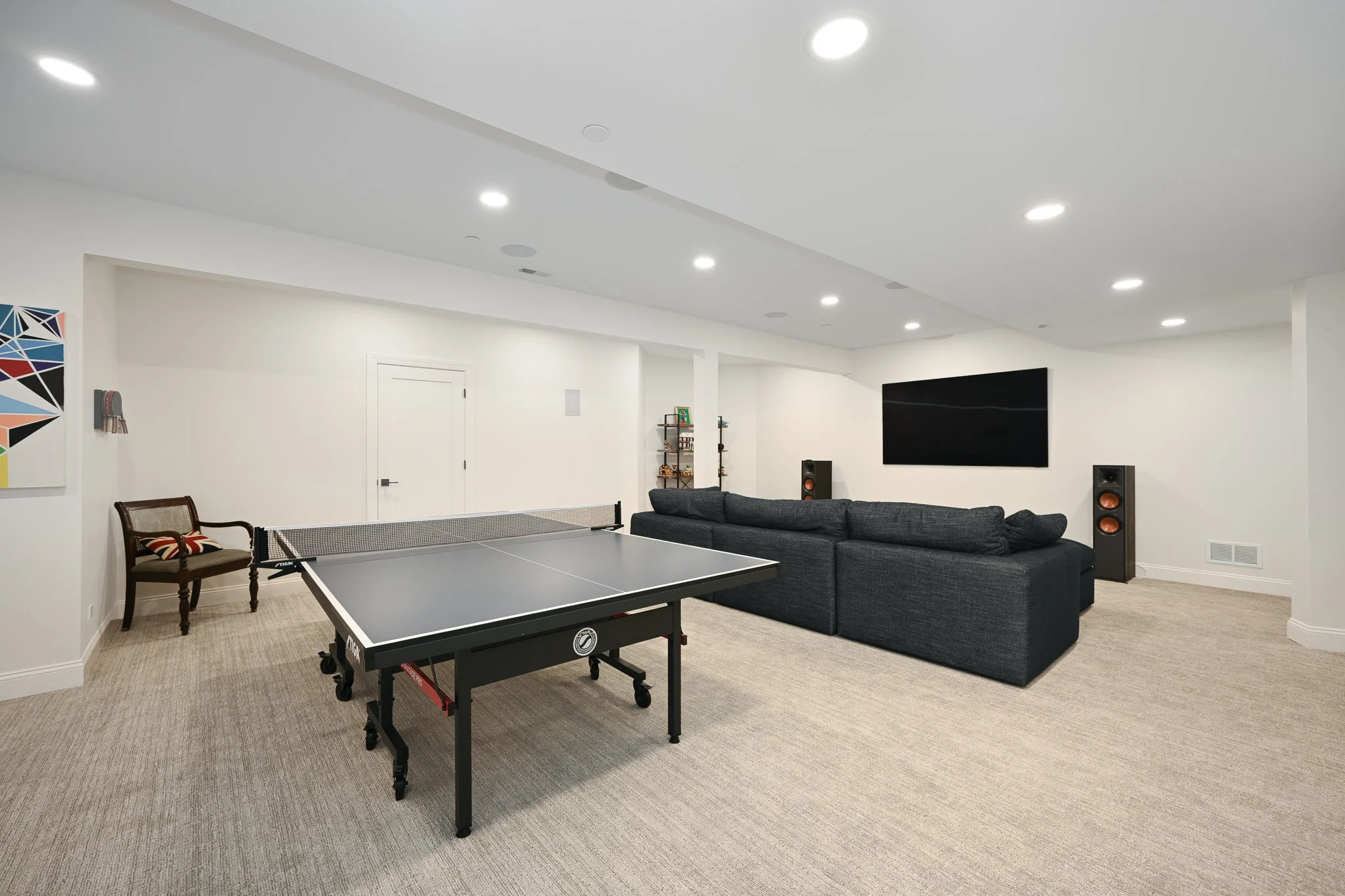 A recreational room with a ping pong table, a large dark gray sectional sofa facing a wall-mounted TV, and stereo speakers on either side of the TV. The room has white walls, recessed ceiling lights, a beige carpet, and minimal decor.