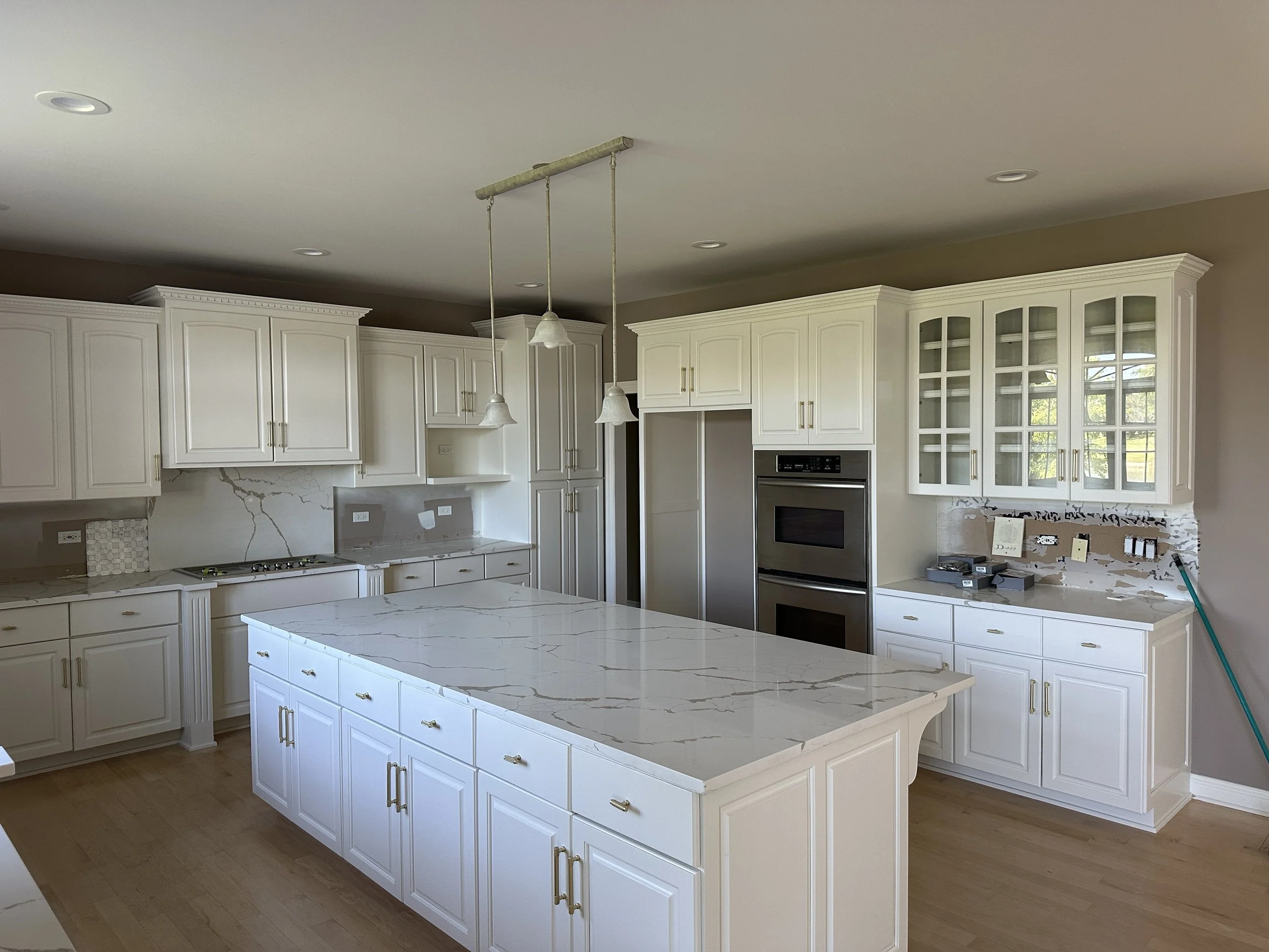 Empty kitchen with white cabinets, marble island, and double ovens, some construction work ongoing.