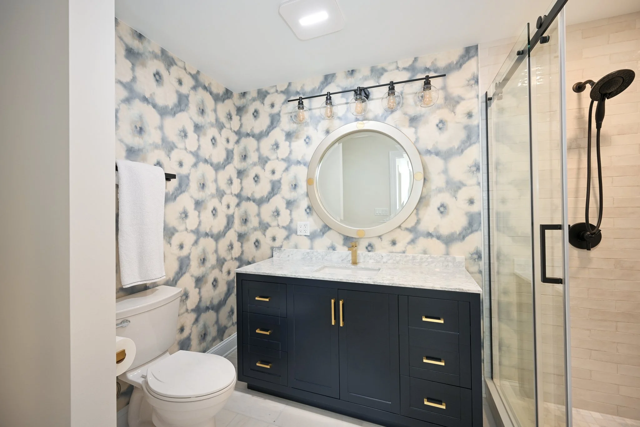 Modern bathroom with patterned blue and white wall art, navy vanity with gold hardware, round mirror, and glass shower with black fixtures.
