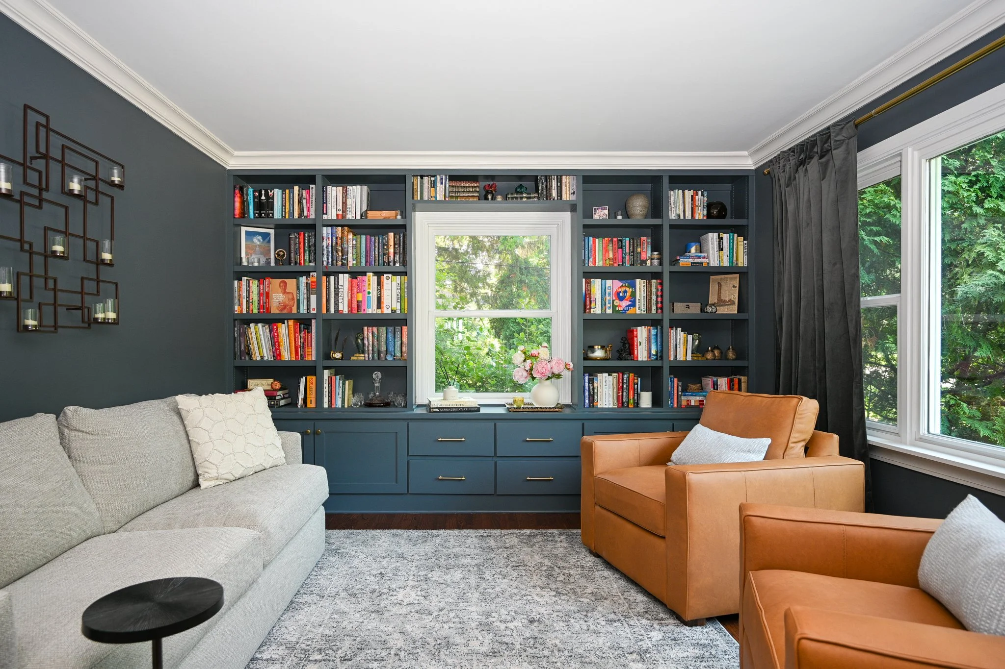 Living room with a built-in dark blue bookshelf filled with books, a window with greenery outside, a beige sofa, two brown armchairs, a gray patterned rug, and decorative wall art.