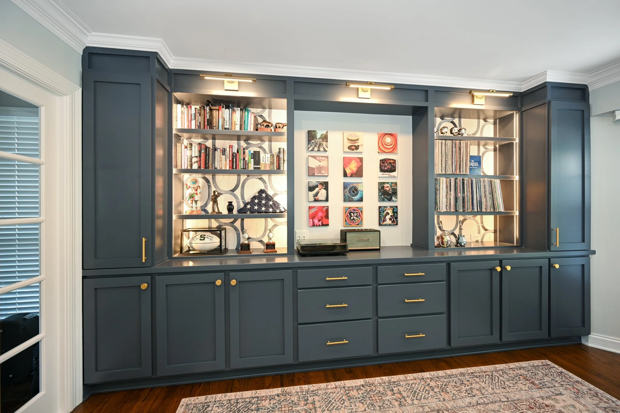 A built-in navy blue bookshelf and cabinet with gold handles, decorated with books, collectibles, and artwork, in a room with hardwood flooring and a patterned area rug.