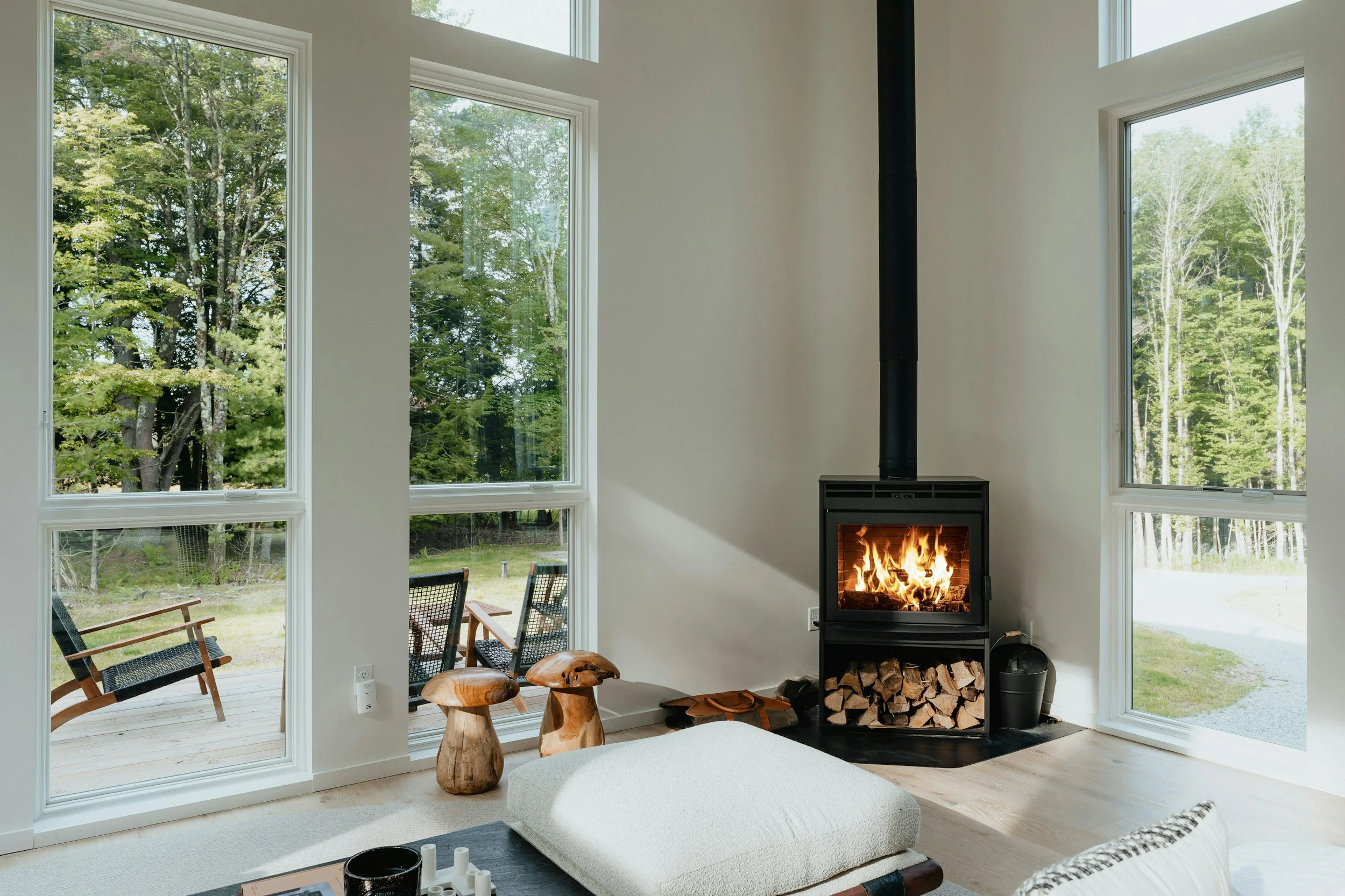 Living room with large windows showing a wooded outdoor scene, a modern fireplace with a wood storage compartment, two wooden stools, a sofa with pillows, and a coffee table with decorative items.