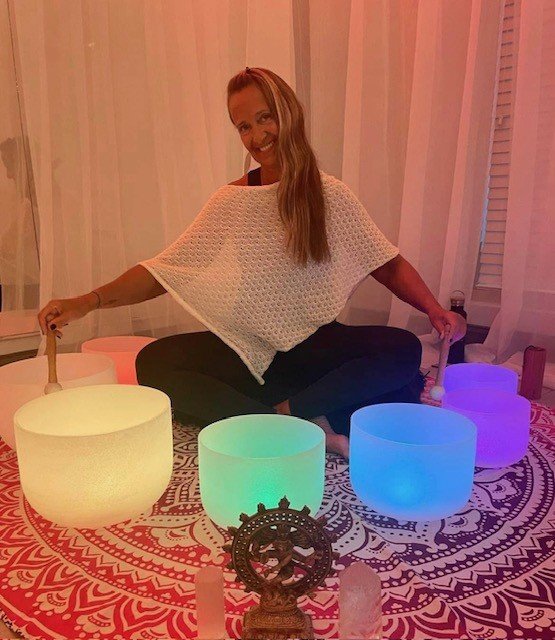Woman sitting cross-legged indoors, surrounded by colorful illuminated crystal singing bowls, with a decorative Ganesha statue and candles in front, on a patterned cloth.