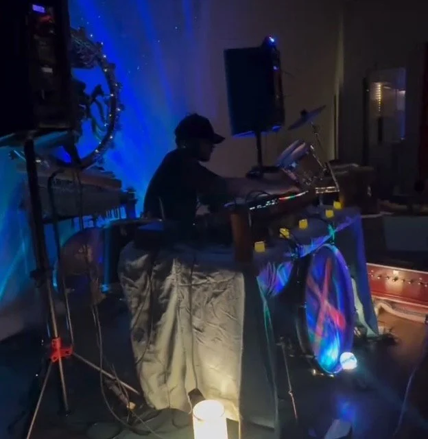 A person wearing a dark cap and shirt performing music DJing at a table with colorful lighting, surrounded by DJ equipment, in a dimly lit room with blue and purple lights.