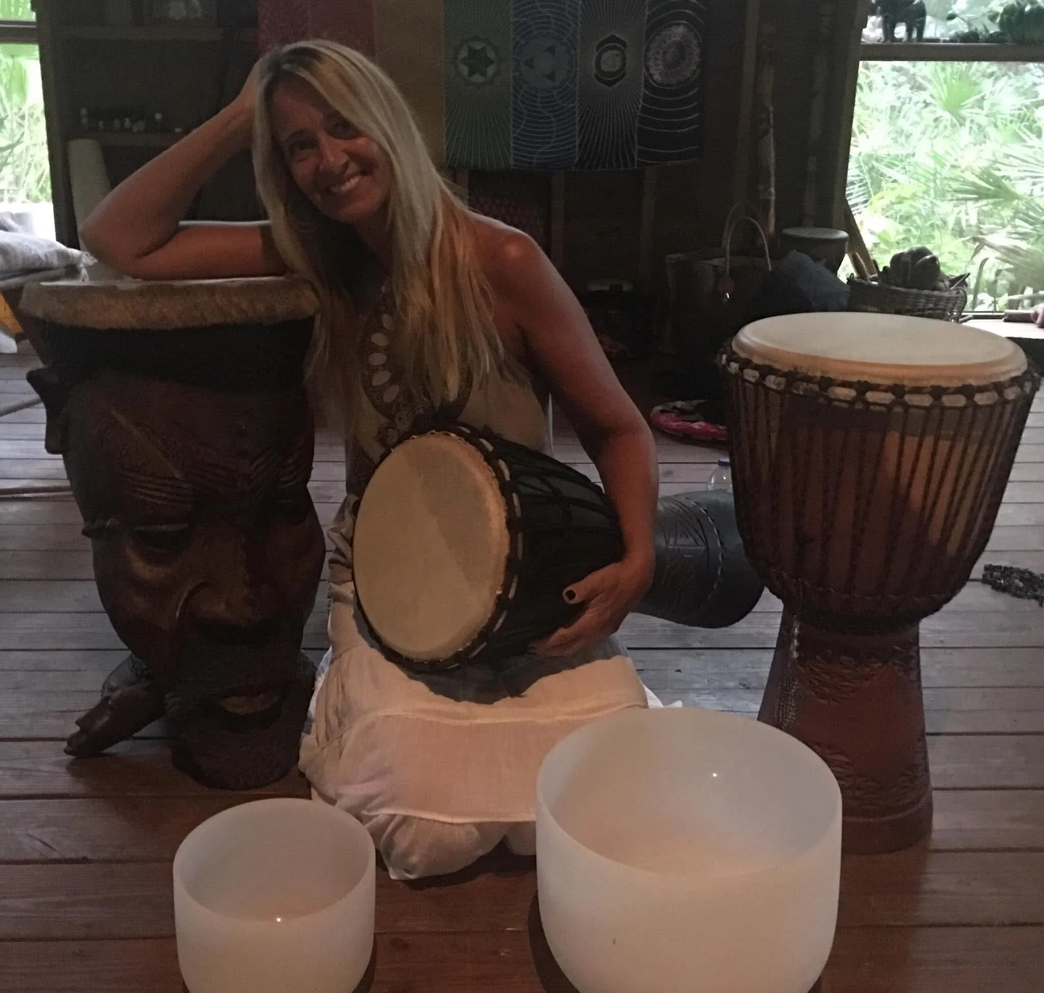 A woman with long blonde hair smiling at the camera, sitting on a wooden floor surrounded by drums and singing bowls, engaging in a musical or creative activity in a room with natural light from a window showing greenery outside.