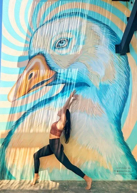 Woman practicing yoga in front of a colorful mural of a bird, with her hands raised above her head and legs in a wide stance.