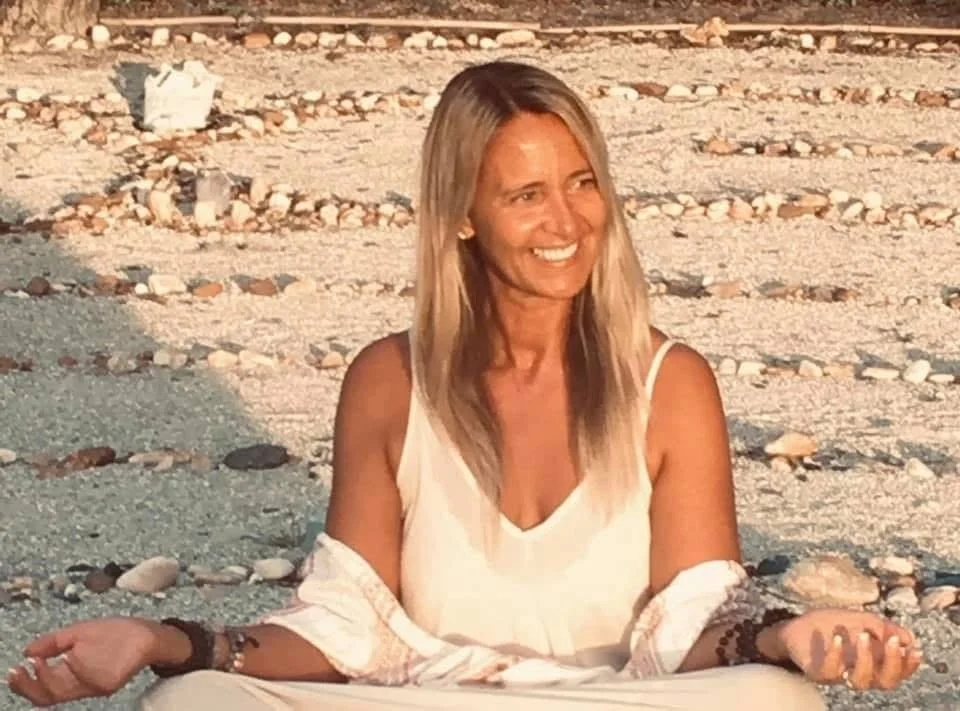 A woman with long blonde hair sitting cross-legged on a pebbly beach, smiling, wearing a white sleeveless top and bracelets, in the warm light of sunset or sunrise.
