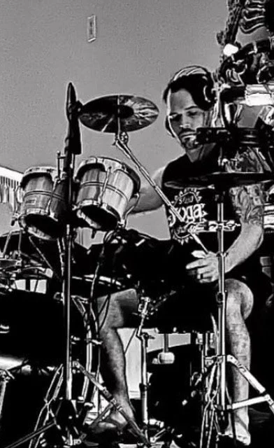 A man playing the drums, surrounded by musical equipment and other items, in a cluttered room.