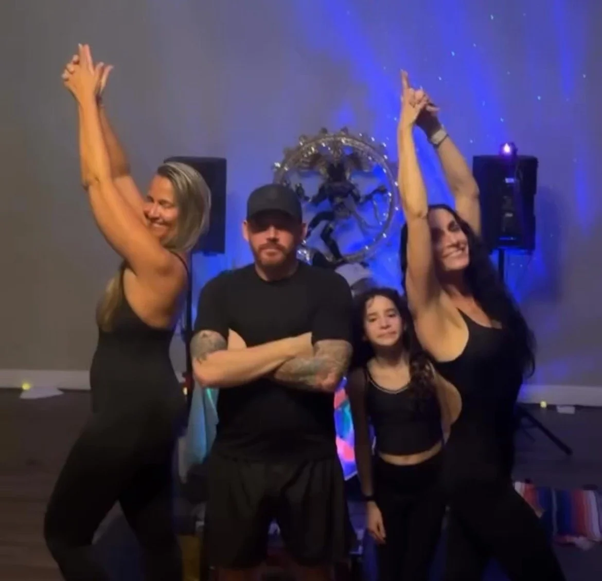 Four people, two women and two children, posing at a party or dance event with a decorative wall art featuring a metallic figure in the background.