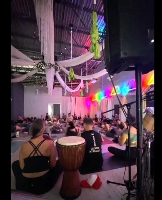 Group of people doing a yoga or meditation session in a decorated indoor space with hanging fabrics and colorful lights.