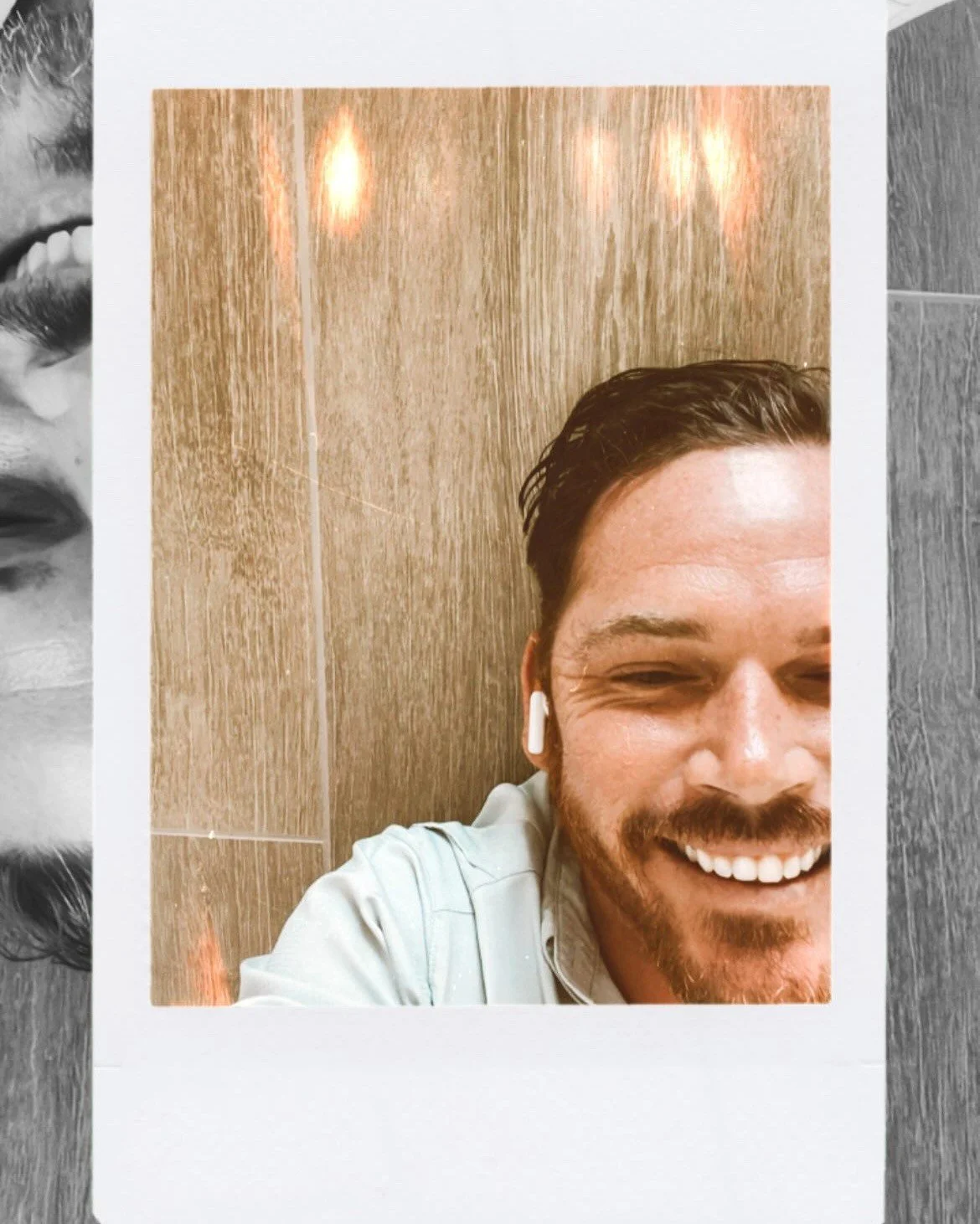 A cheerful man smiling with white teeth, wearing white wireless earbuds and a light-colored shirt, taking a selfie in front of a wooden textured background.