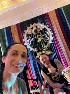 Two women taking a selfie in front of a colorful, striped fabric backdrop with a large decorative Ganesha statue.