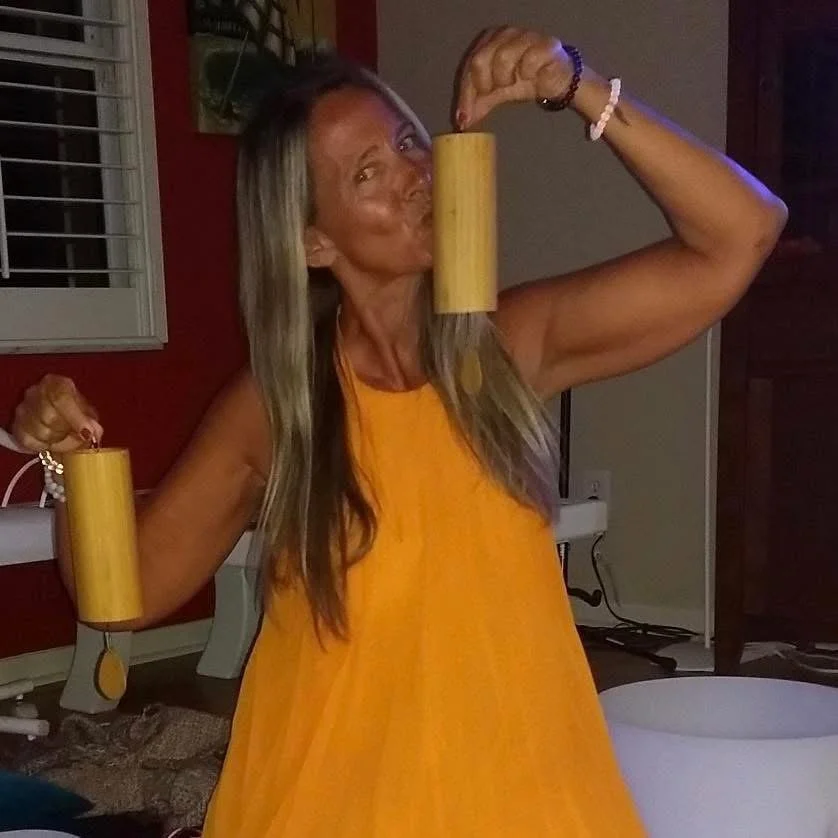 A woman in a yellow dress flexing her muscles while holding two wooden percussion instruments, one in each hand, in a dimly lit room.