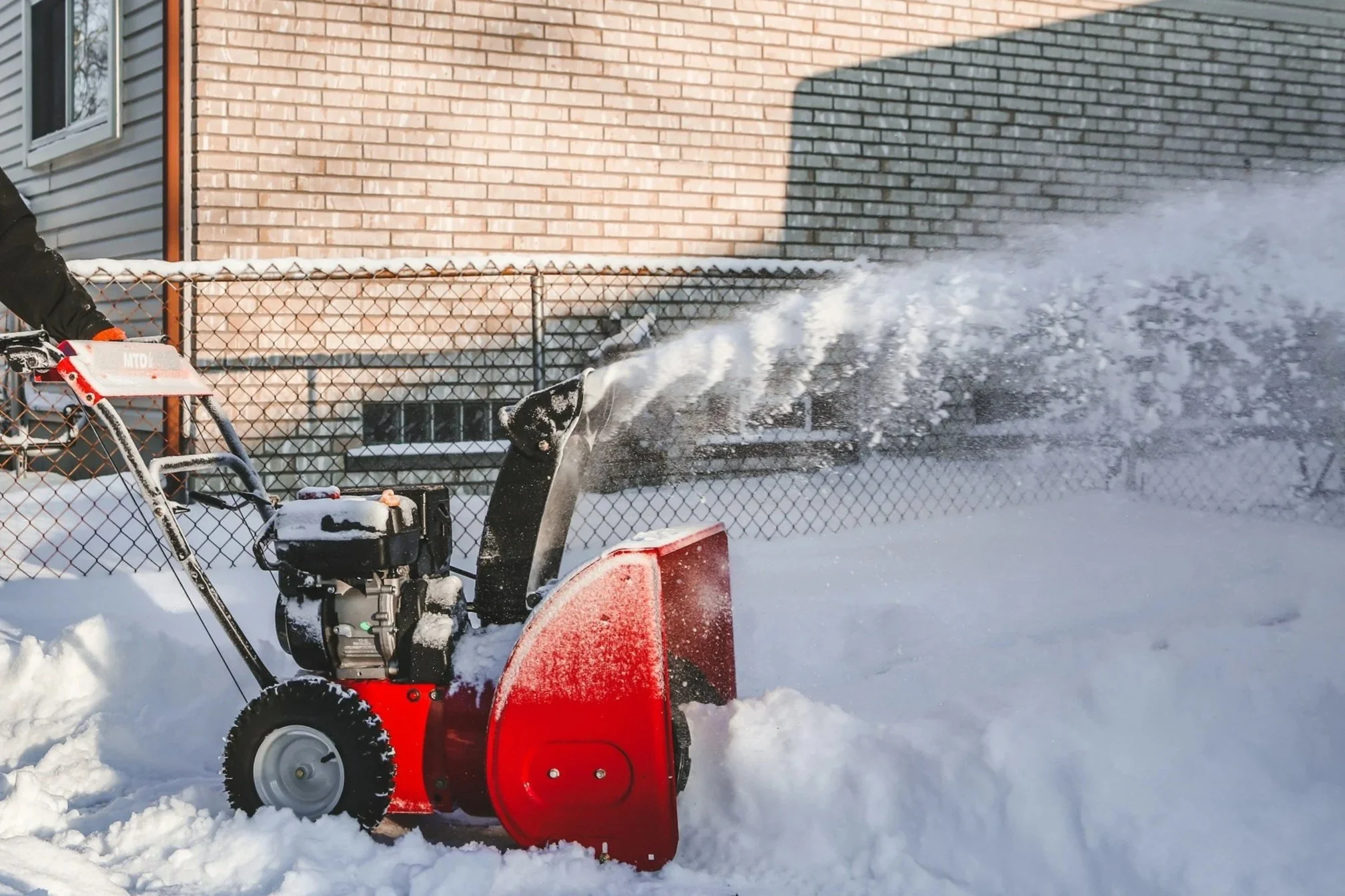 A person using a red snow blower to clear snow in a backyard, with snow flying through the air.