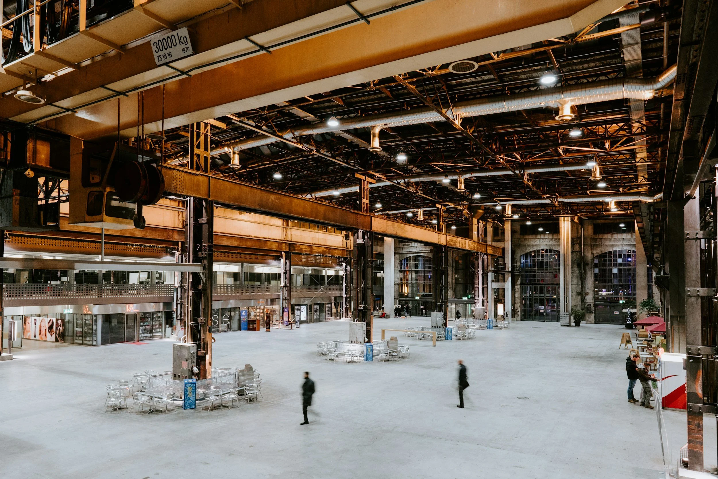 Large, empty industrial-style indoor space with high ceilings, metal beams, and overhead lighting. Four people are walking or talking, and one counter with two people is visible on the right side.