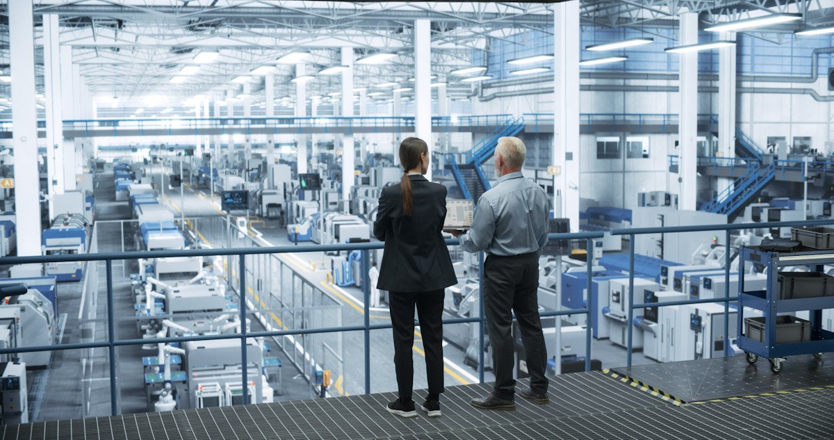 Two professionals, a woman and a man, stand on an observation balcony overlooking a large, modern factory or warehouse filled with automated machinery and conveyor belts.
